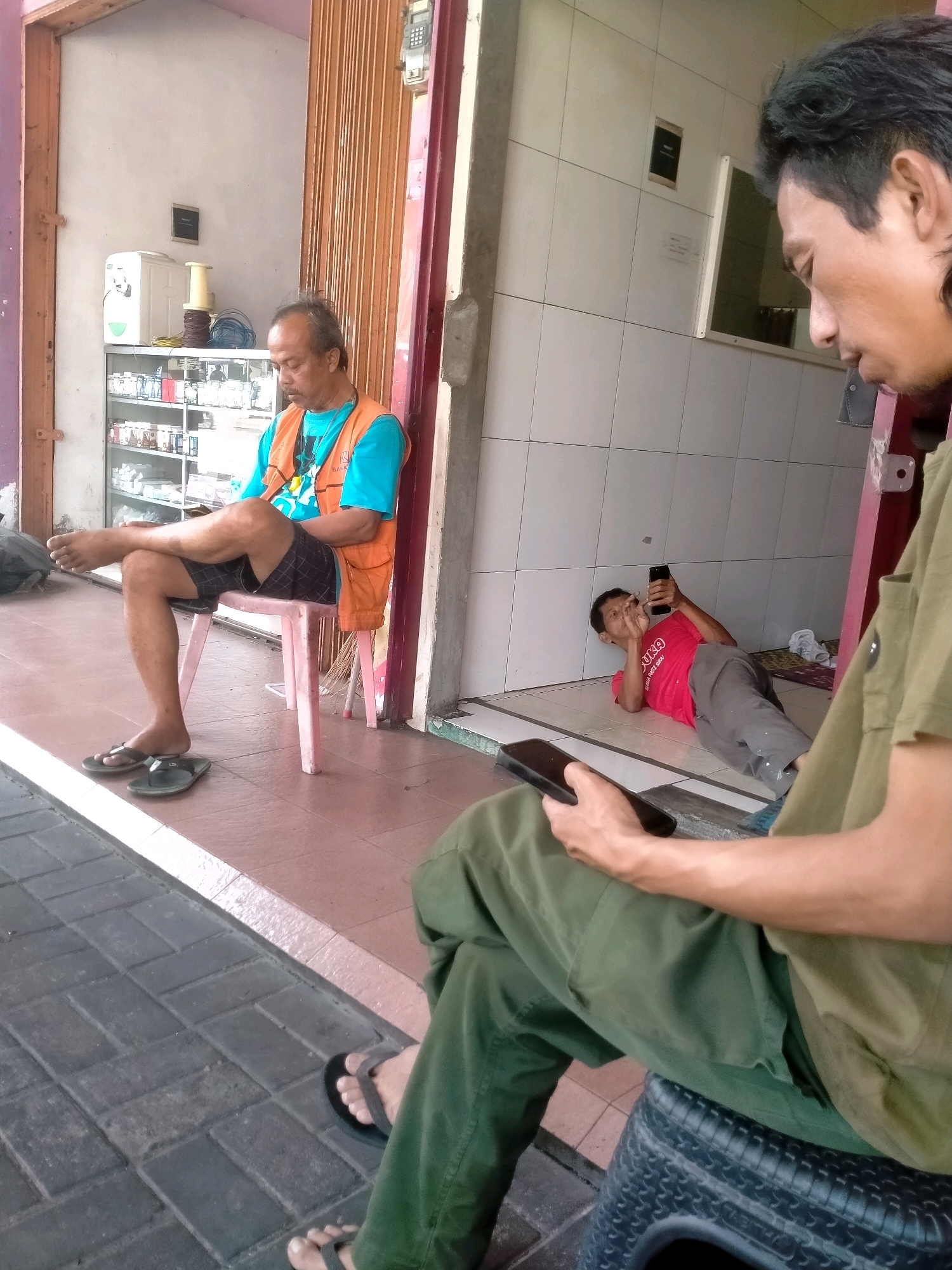 The image depicts an outdoor scene in front of a shop in Kasihan, Indonesia. In the foreground, a person wearing a green shirt and pants is seated on a black tire. To their left, another person wearing a bright blue shirt and an orange vest is sitting on a pink chair with their legs crossed, looking down. In the background, a third person in a red t-shirt and grey pants is lying on the floor, looking at a phone. The shop appears to be a retail establishment, with shelves stocked with items visible through the open doorway. The ground is paved with dark gray brickwork. The lighting suggests it is daytime, likely with overcast skies or partial shade.