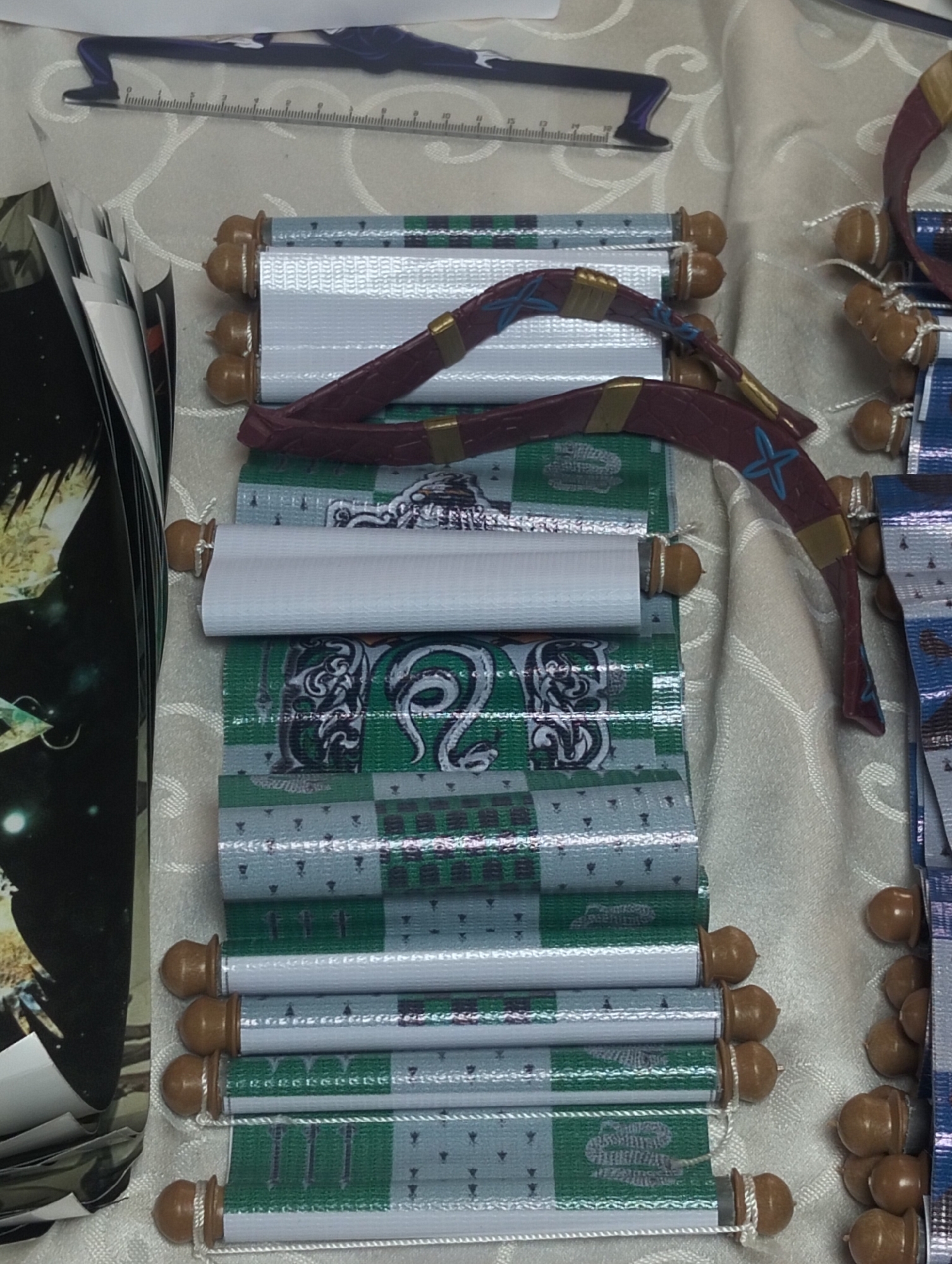 The image shows a collection of decorative scrolls, likely souvenirs or merchandise, arranged vertically on a textured white cloth. The scrolls are cylindrical, with a light-colored paper or fabric rolled around a core, and topped with small, rounded brown finials at each end. Many of the scrolls are decorated with repeating patterns in green and silver or white. Some of these patterns appear to be stylized snakes or crests. One scroll has a dark red ribbon tied around it, embellished with gold and blue accents. To the left of the scrolls, a stack of glossy printed sheets or cards with a dark background and golden or yellow imagery is partially visible. A ruler is visible in the background, along with a silhouette of a figure.
This image does not provide any information that would allow for the determination of its location in the city of Dhaka, Bangladesh, or any other specific geographic location.