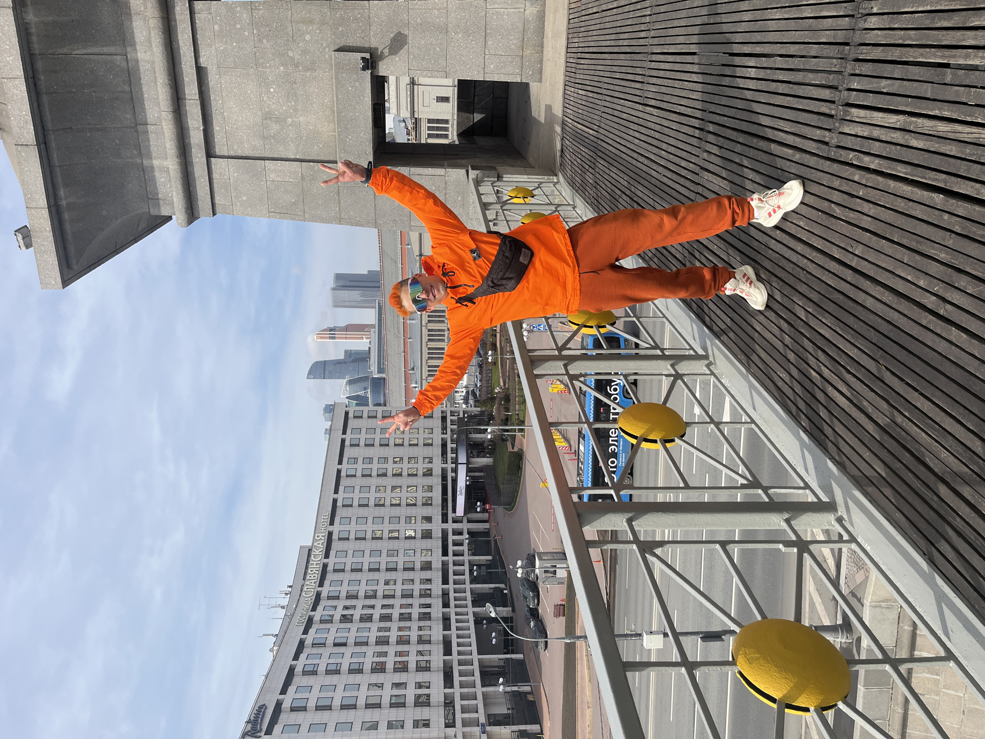 A man in an orange tracksuit strikes a celebratory pose on a bridge, with the Radisson Slavyanskaya Hotel and skyscrapers in the background, situating the scene in Moscow, Russia. He is positioned at an angle, facing towards the city. His arms are outstretched, with his hands making the "peace" sign. The man's attire includes white sneakers with red accents, a black waist pack, and colorful sunglasses. He is standing on a grey metal railing with yellow decorations. The background features a light blue sky with scattered clouds.