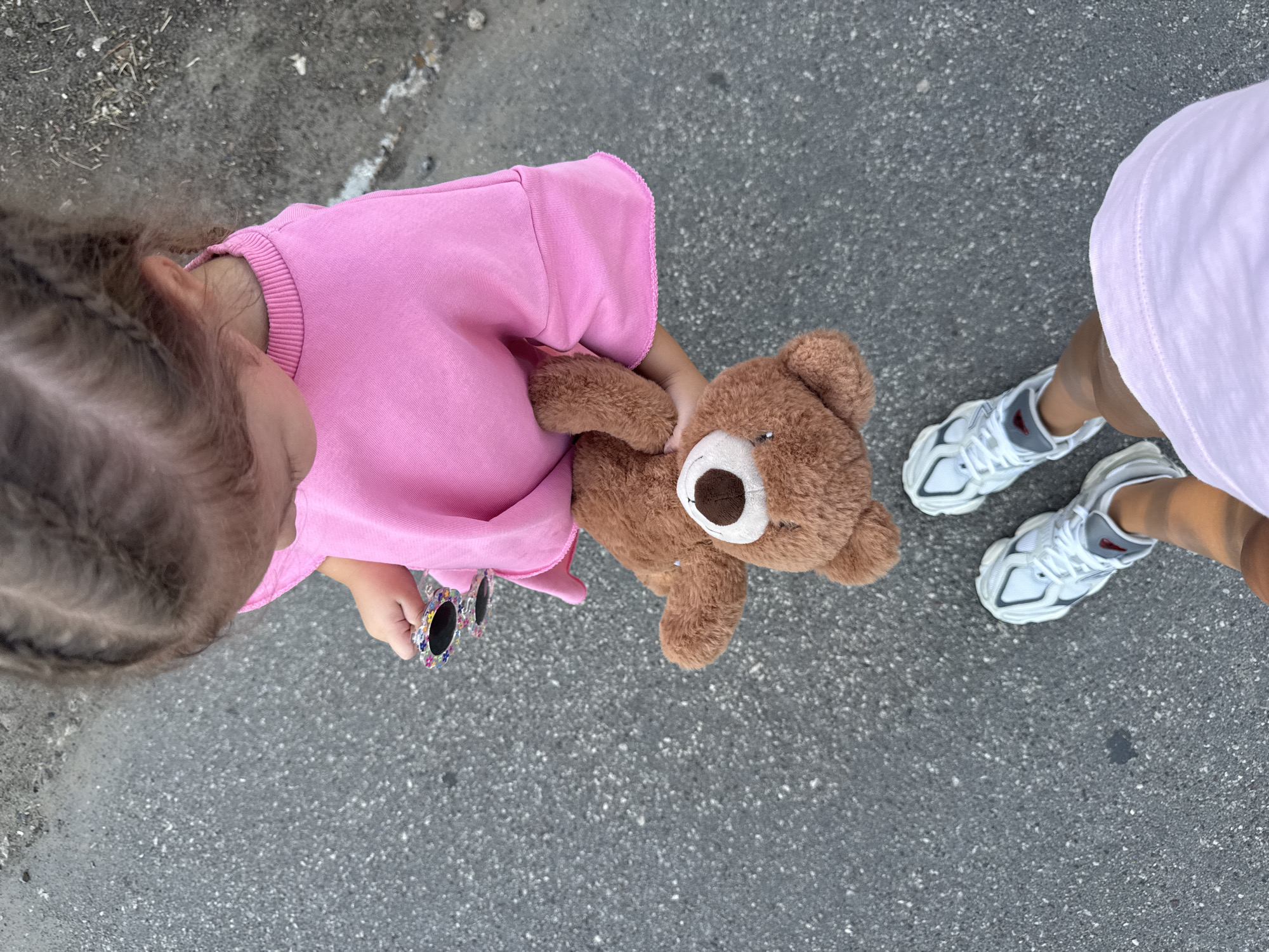 In a top-down perspective, a young child wearing a pink sweatshirt and white sneakers is holding a brown teddy bear and a pair of decorative sunglasses. The child's hair is styled in braids. The scene appears to be outdoors on an asphalt surface, possibly a sidewalk or street. The ground has a textured appearance with small pebbles and debris. The lower portion of the image shows another person's legs and feet, also wearing white sneakers and a light pink garment. The overall lighting suggests daytime. This image does not contain enough information to determine a specific location in Chișinău, Moldova.