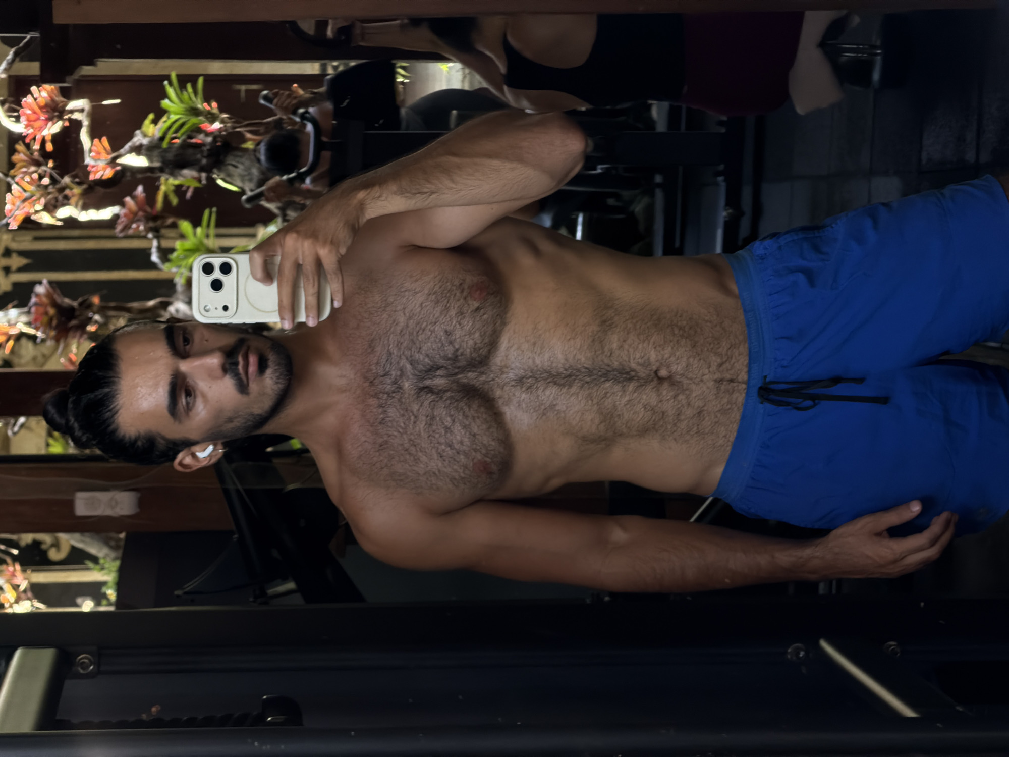 A mirror selfie captures a man, oriented horizontally across the frame, in what appears to be a gym or a home setting in North Kuta, Indonesia. The man has dark hair tied in a bun, a beard, and a mustache. He wears a white earbud in his right ear. He is shirtless, revealing a muscular, hairy chest and arms, and is wearing blue athletic shorts. In his right hand, he holds a light-colored iPhone with three camera lenses, used to take the picture.
The background, visible through the mirror's reflection, features dark wooden structures and vibrant green and orange plants, suggesting a tropical and possibly outdoor or semi-outdoor environment. Faint reflections of dark objects, resembling gym equipment or furniture, and another person wearing a dark top, are also discernible. The lighting is bright and diffused, indicating it is daytime. The man maintains a neutral expression. No visible text is present in the image.