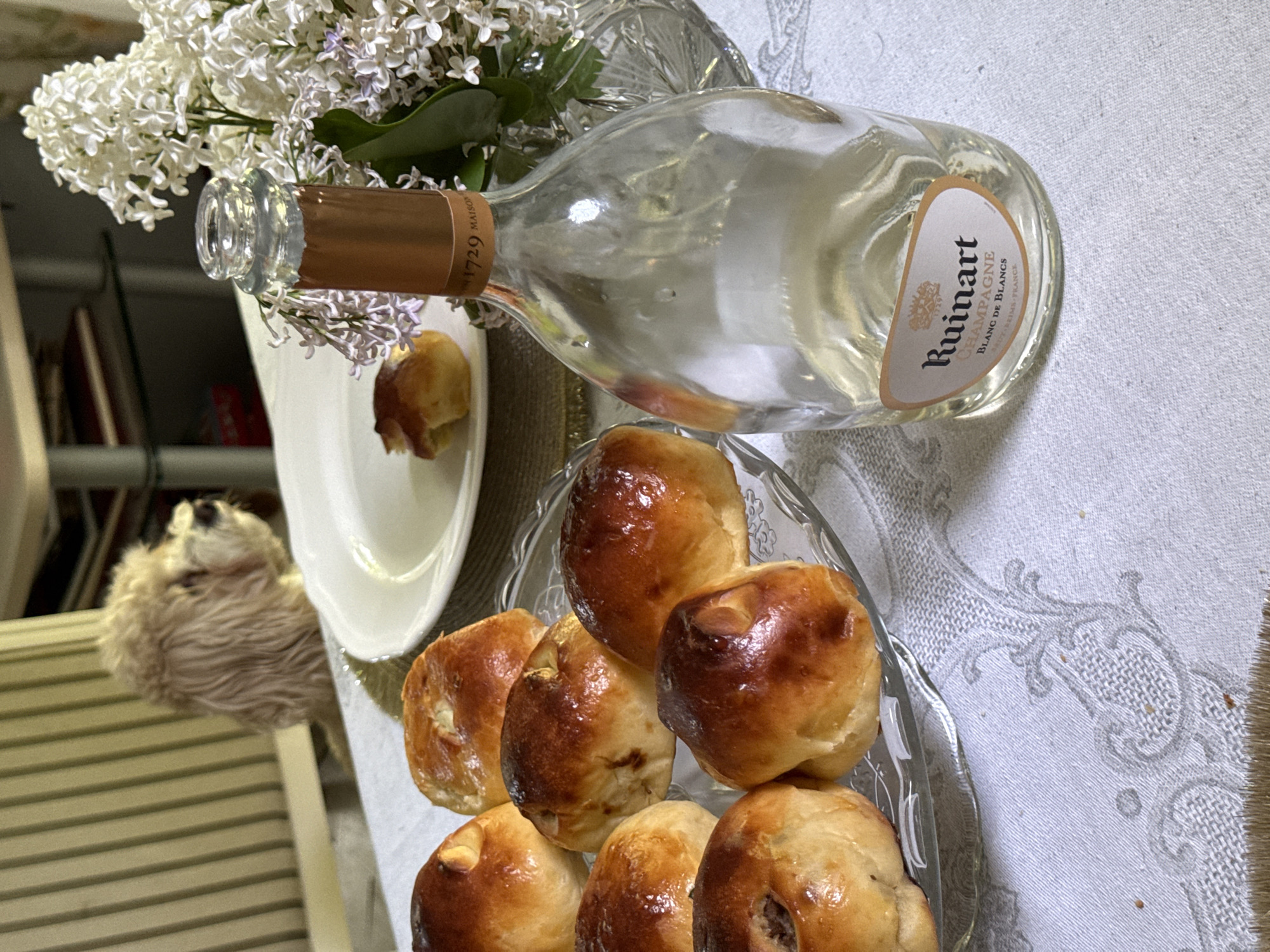 The image depicts a still life arrangement on a table. A bottle of Ruinart Blanc de Blancs champagne is positioned horizontally, near a plate with a partially eaten pastry. In the foreground is a glass dish containing several small, baked buns or pastries. White lilac flowers in a vase are visible in the upper left corner. A small, fluffy dog is seated on a chair in the background. The scene appears to be indoors, on a table covered with a patterned tablecloth. There is no evidence to suggest it is located in Sapronovo, Russia.