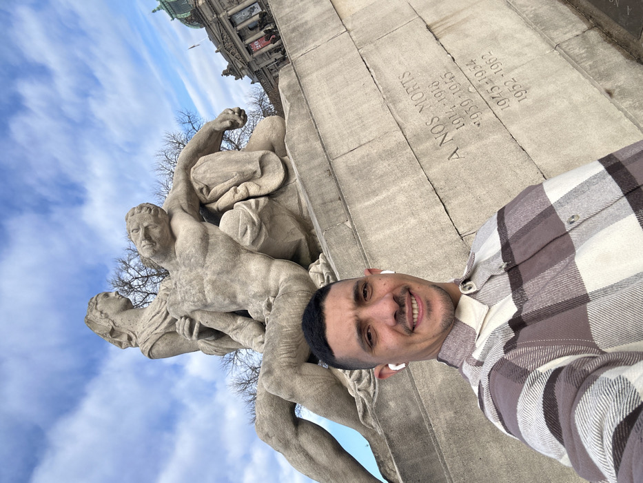 An outdoor, low-angle selfie depicts a male subject standing in front of the Monument aux Morts (War Memorial) located at Place de la République in Strasbourg, France. The man, with short dark hair and a light mustache, wears a brown, white, and tan plaid shirt and white wireless earbuds, smiling at the camera. Directly behind him, the large stone monument features an allegorical female figure supporting a deceased male soldier. The monument's base is inscribed with "A NOS MORTS" and lists the dates "1914-1918", "1939-1945", "1945-1954", and "1952-1962". In the background, the distinctive green dome and classical architecture of the Palais du Rhin are visible against a blue sky with scattered white clouds. Bare tree branches are also present.