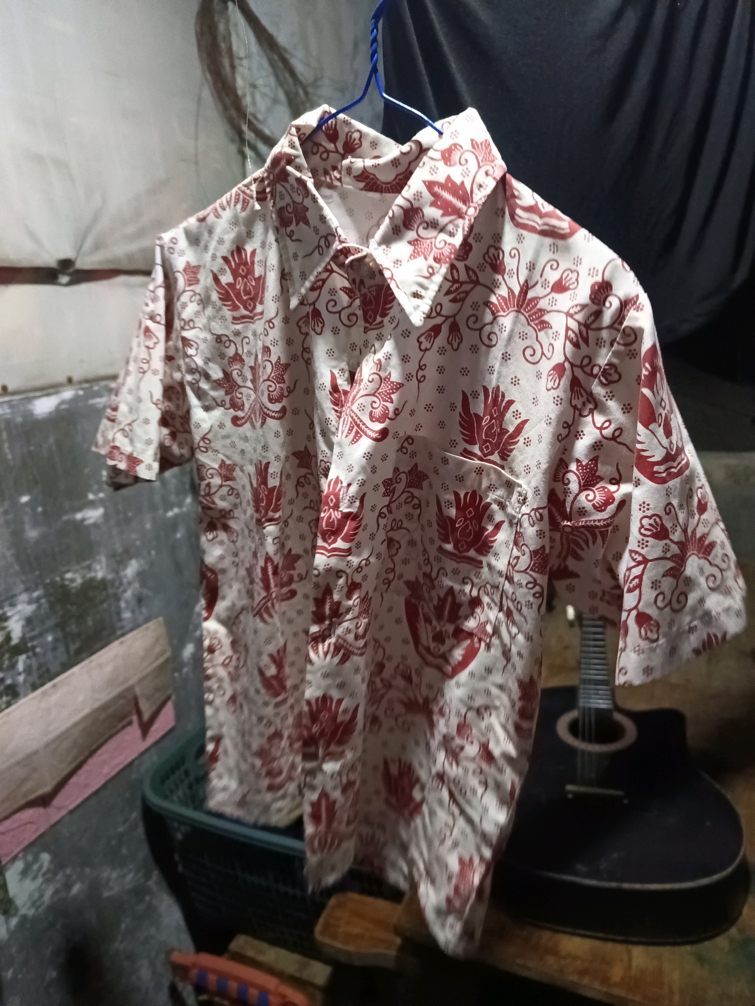 A short-sleeved, collared shirt with a batik pattern of red birds and floral motifs on a cream background hangs from a blue plastic hanger. The shirt is draped over a dark-colored acoustic guitar. In the background, a dark cloth hangs, and to the left, a gray concrete wall is visible. The setting appears to be an indoor space, possibly a workshop or storage area, with a basket and wooden objects in the foreground. The lighting is dim, creating shadows and highlights. The scene is likely located in a residential or commercial area within the city of South Jakarta, Indonesia.