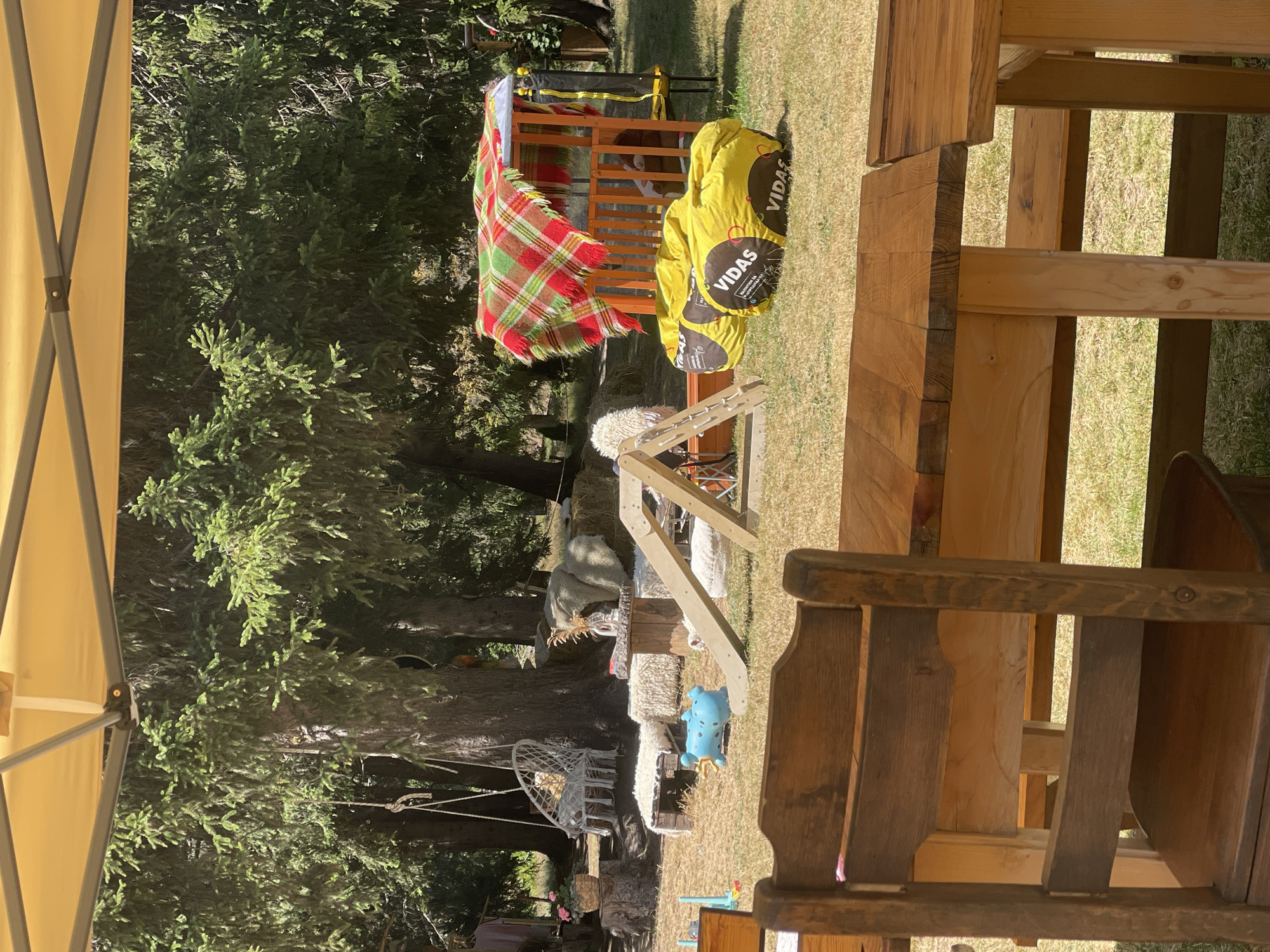 The image shows an outdoor seating area under a yellow canopy, with a wooden table and chairs in the foreground. In the background, there is a playground with a slide, yellow beanbags, and a small playhouse covered with a checkered blanket. There are also trees, hay bales, and a white hanging chair visible. The location is identified as the city of Smolyan, Bulgaria. The scene appears to be set up for a family-friendly outdoor event or play area.