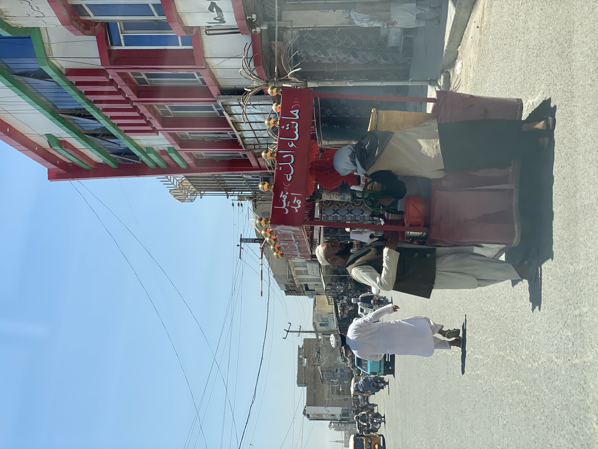 This image captures a street scene in Kandahar, Afghanistan, on a bright, sunny day. A man in traditional clothing, including a white cap and a vest over a long tunic, is standing near a red vendor cart. The cart has Arabic script on a red sign above it and is decorated with golden orbs. Another person, partially obscured, is operating a green mechanism on the cart, possibly preparing a drink. In the background, a multi-story building with red and green accents dominates the right side of the frame, featuring numerous windows. The street is paved and appears to be a busy thoroughfare, with various vehicles, including motorcycles and a three-wheeled auto-rickshaw, moving along it. Several other individuals are visible on the street, some walking and others on motorcycles. The sky is clear and blue with a few overhead wires. The overall atmosphere suggests a typical urban environment with commercial activity.