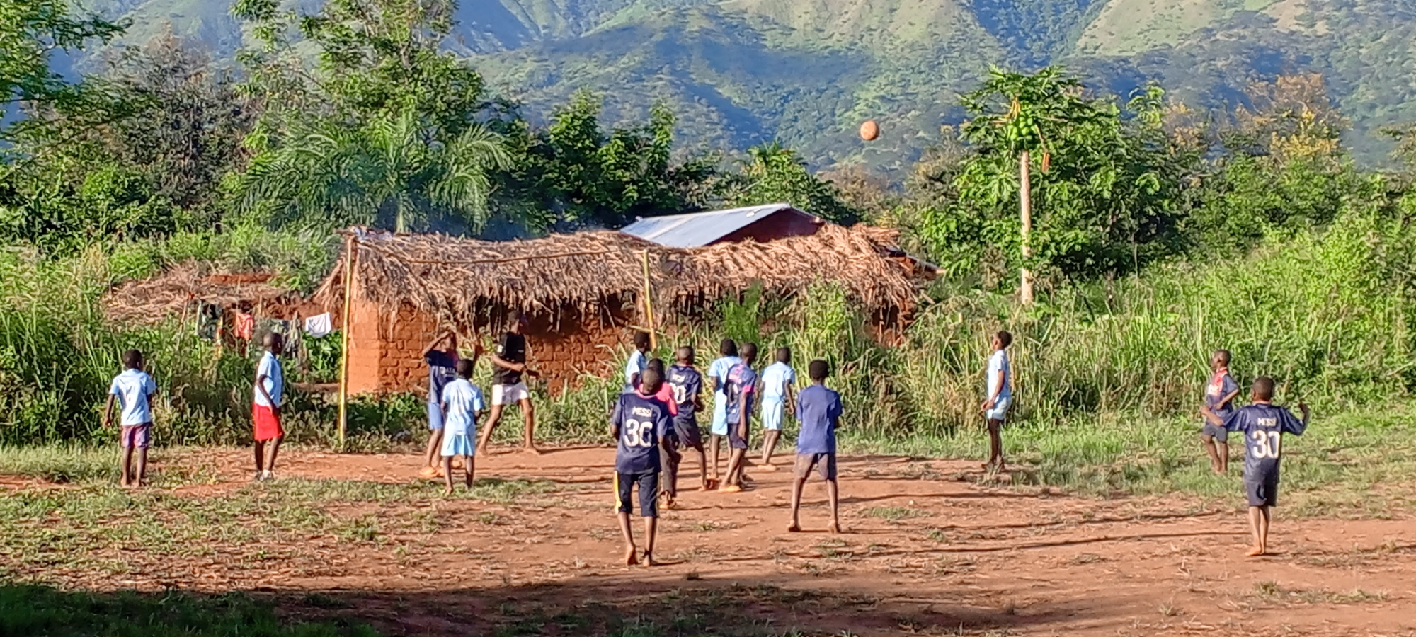 A group of approximately a dozen children are playing on a dirt field in Ngalula, Democratic Republic of the Congo. The children are of varying heights and are wearing an assortment of clothing, including t-shirts, shorts, and several soccer jerseys with the name "Messi" and the number 30 printed on the backs. A ball is in the air above the children, seemingly mid-play. In the background, there is a small mud hut with a thatched roof and laundry hanging outside. Lush green vegetation surrounds the playing area. In the distance, mountains are visible. The light suggests it is either early morning or late afternoon.
