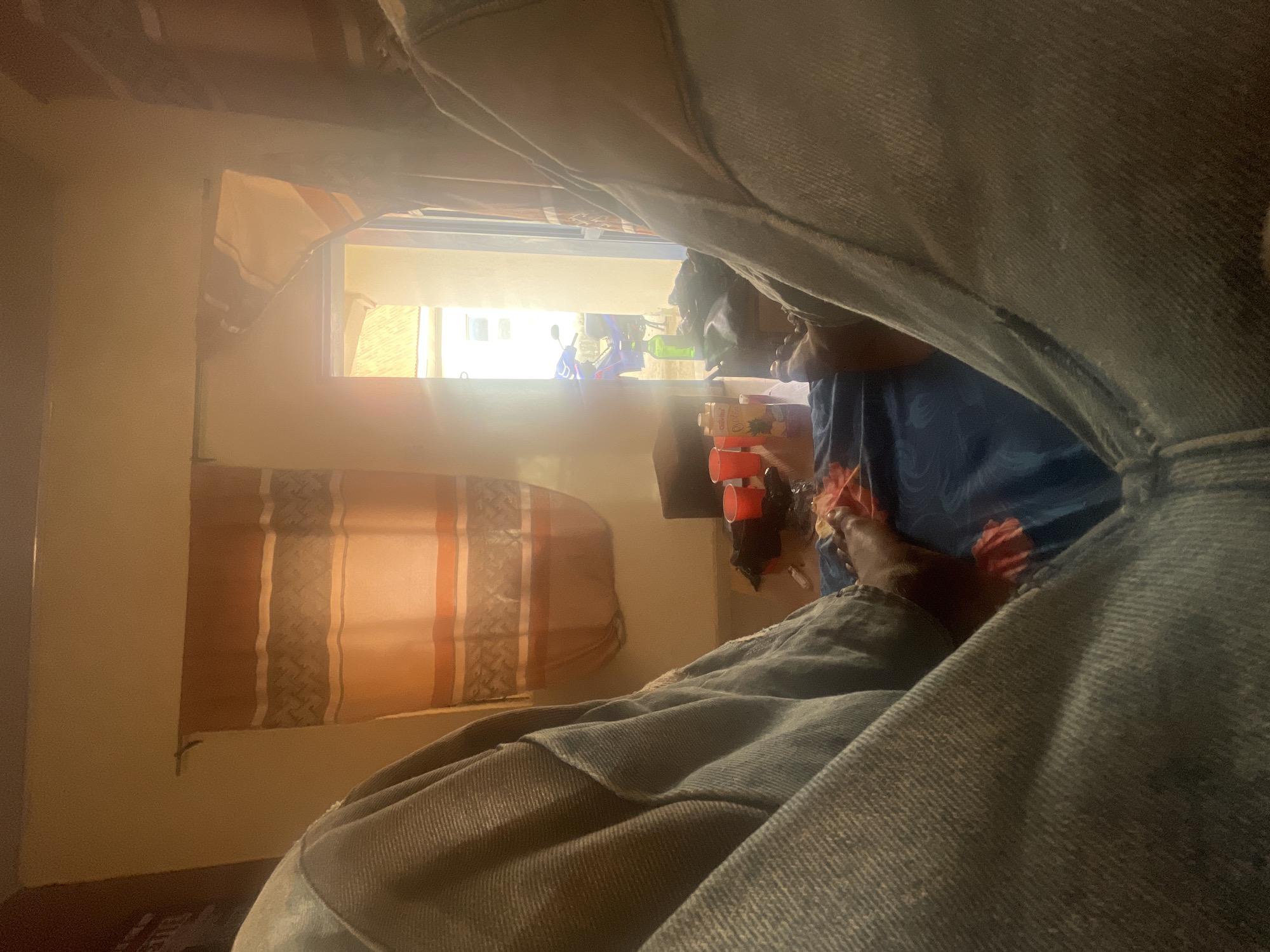 The image captures an intimate indoor scene from a very low, upward-looking perspective, dominated by folds of light-colored denim fabric, likely jeans, belonging to the person taking the photograph. A dark-skinned foot, possibly wearing a sandal, is visible in the lower right, confirming the presence of an individual lying down or resting.
The primary light source is a brightly illuminated window, revealing a glimpse of the outside or another brightly lit room beyond. To the left of the window, a curtain or blind with horizontal stripes in brown, orange, and cream tones is visible, along with another piece of patterned fabric hanging above. The interior walls appear to be a light, neutral color.
On the right side, beneath the window, several objects are stacked, including a carton clearly labeled "JUICE" and "PINEAPPLE," along with some red cups. Through the overexposed window, blurry figures, one wearing blue, and possibly a blue and white scooter or motorcycle, can be vaguely discerned, indicating activity in the distant background.
The intense sunlight suggests it is daytime. The setting appears to be residential or domestic, given the presence of curtains and household items. The overall mood is quiet and personal, capturing a moment of repose from an unusual, first-person vantage point in what is understood to be Jos, Nigeria.