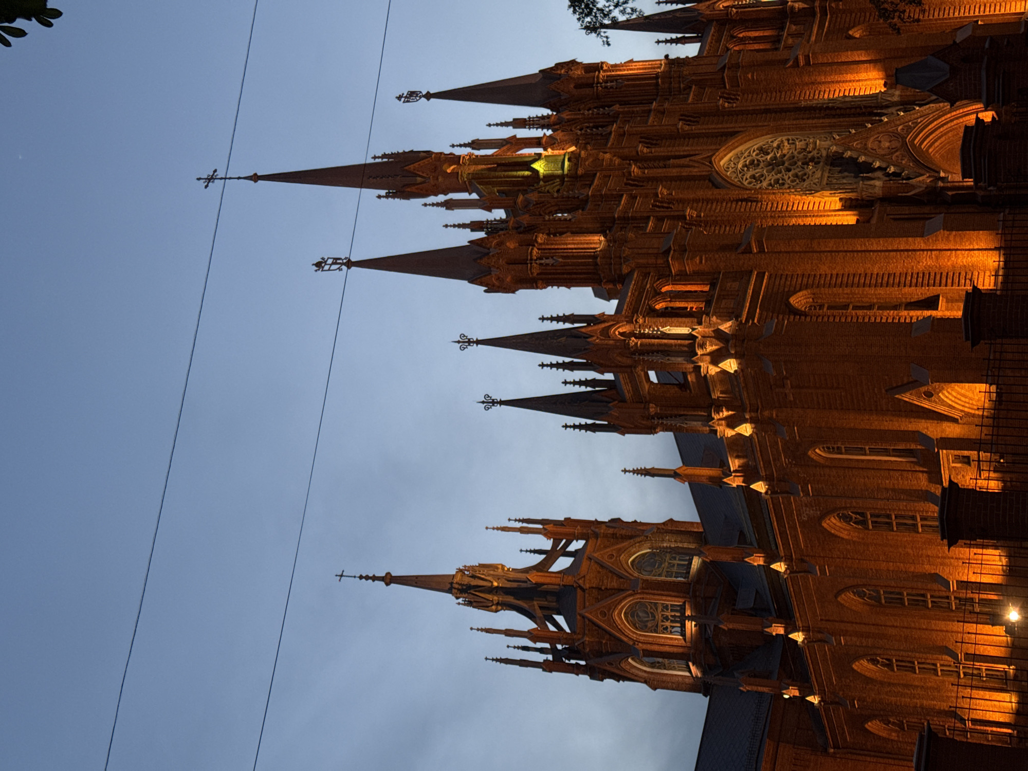 The image depicts the Catholic Cathedral of the Immaculate Conception in Moscow, Russia, at twilight. The facade is illuminated with a warm, golden light, accentuating the Gothic Revival architectural details such as spires, arches, and intricate ornamentation. The sky is a pale blue, indicative of dusk. Power lines cut across the upper left portion of the frame. The building appears to be constructed of red brick. There are no people visible in the image. The image is oriented sideways.