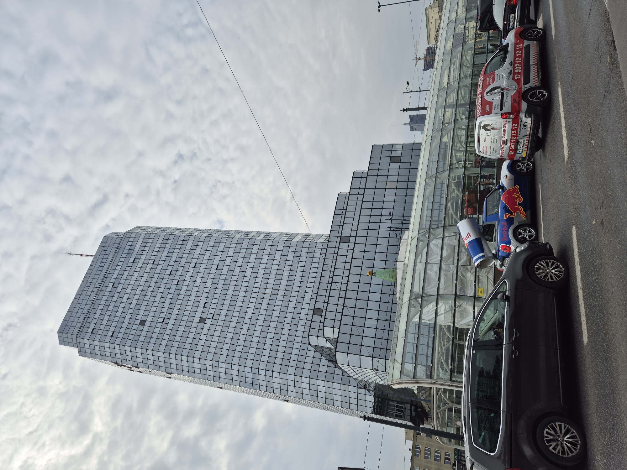 The image captures a street scene in Warsaw, Poland, featuring the Rondo 1 skyscraper, a modern high-rise with a glass facade. The building is set against a cloudy sky. In the foreground, there are several parked vehicles on the side of the road, including a dark-colored car, a Red Bull-branded car with large Red Bull can props, and a van with locksmith services advertised on its sides. The van has contact information including the phone number 507 12 12 12 and license plate WH 4708R. Other vehicles are visible further along the street. A glassy pedestrian overpass/structure is adjacent to the skyscraper. Power lines stretch across the sky. Overall, the image presents a mix of urban architecture, advertisement, and street life in a major city.