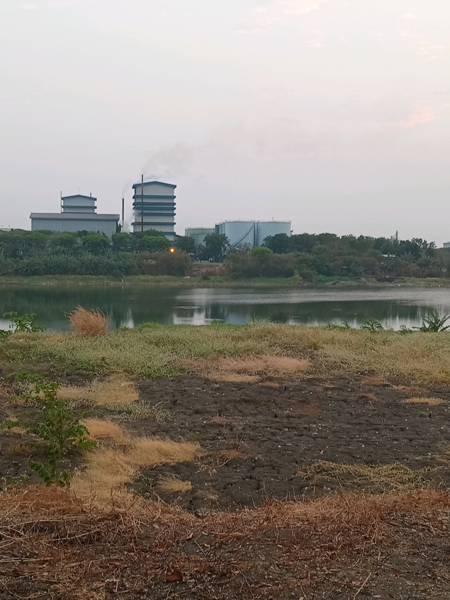 An industrial facility is depicted in the distance across a body of water in Karangturi, Indonesia. The facility features several multi-story buildings, including one with a prominent smokestack emitting a plume of smoke into the overcast sky. Adjacent to these structures are large, cylindrical tanks, likely for storage. The foreground of the image shows a dry, barren landscape with patches of withered grass and scattered debris. A small green shrub is visible in the lower-left quadrant. The body of water in the middle ground reflects the muted tones of the sky and the industrial structures. The overall impression is one of an industrial zone situated near a natural body of water, with a slightly hazy atmosphere.