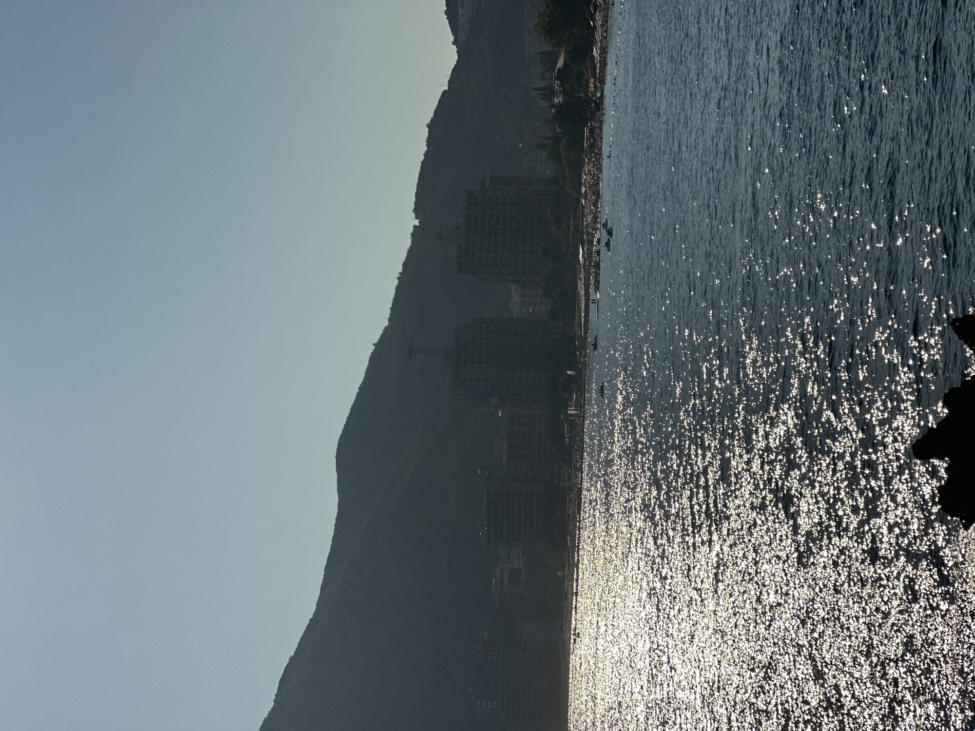 The image shows a view of the coastline in Budva, Montenegro, during daylight. The foreground is dominated by the dark blue sea, with the surface shimmering intensely due to sunlight reflecting off the waves. In the midground, a crowded beach is visible, lined with numerous white umbrellas and sunbathers. Beyond the beach, a line of buildings, including several multi-story structures, some appearing to be under construction, form the urban landscape. Behind the buildings, a large, dark, tree-covered mountain slopes upwards, its silhouette stark against the clear, pale blue sky. The overall scene depicts a sunny day at a popular coastal resort.