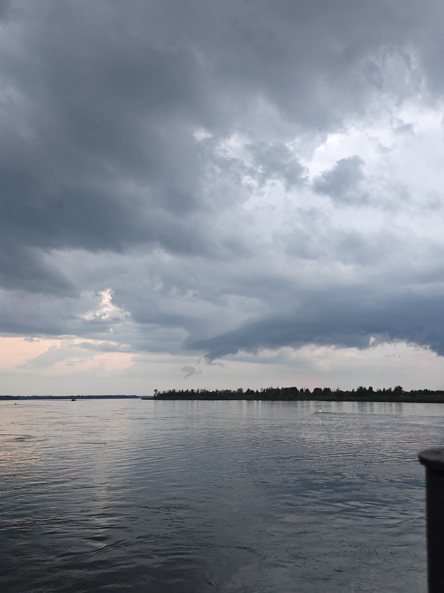 A wide shot captures a body of water under a heavily clouded sky. The sky is filled with dark, imposing storm clouds, with a few lighter patches hinting at the sky beyond. The water's surface is rippled, reflecting the muted tones of the sky. In the distance, a thin strip of land is visible, densely forested with trees. Two small boats are present on the water: one closer to the left bank, and another further out, near the right bank. A dark railing or edge of a structure is visible in the lower right corner of the image. The location is identified as Kazachinskoye, Russia.
