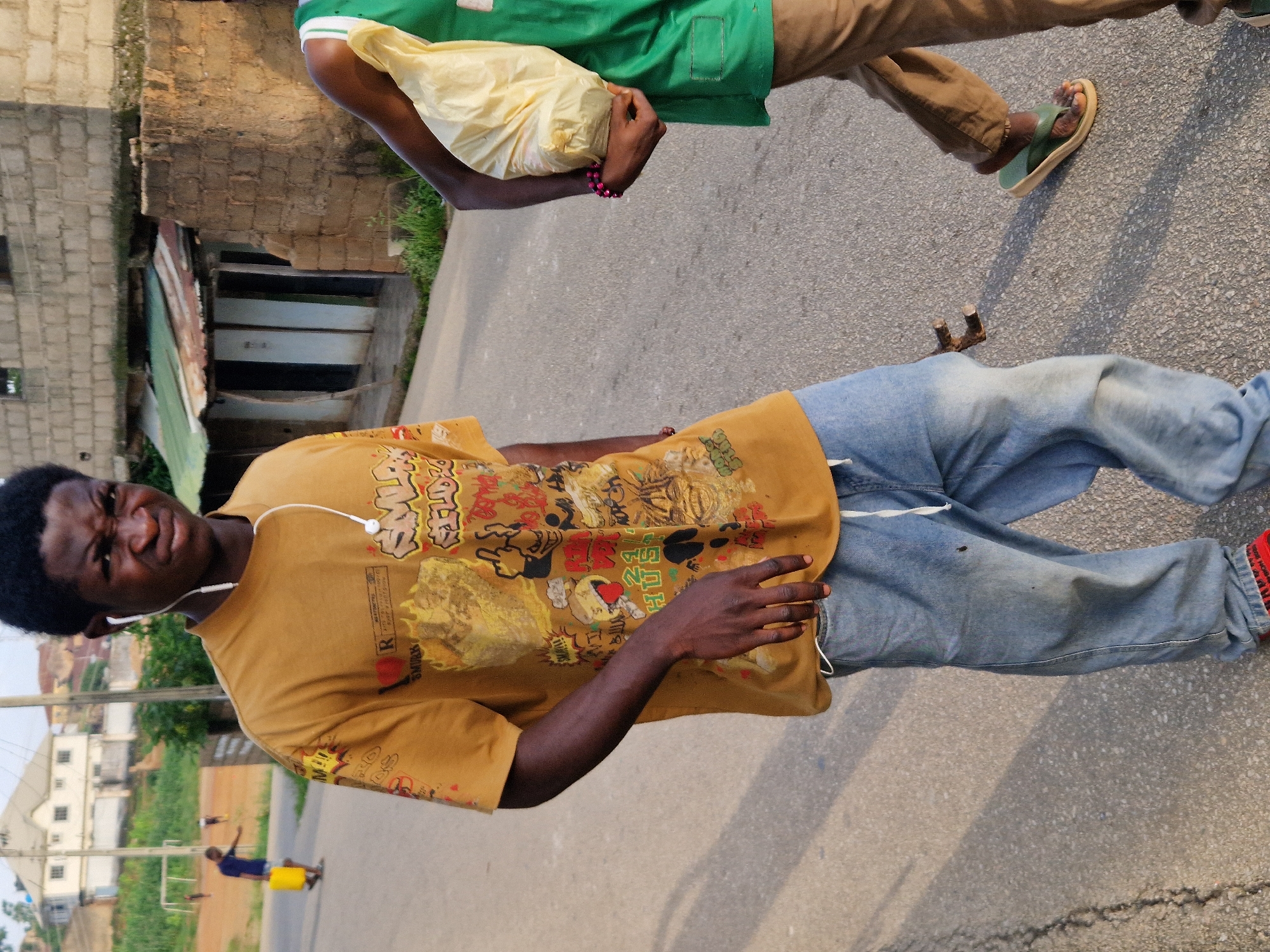 The image shows a young Black male walking on a paved street in Kabong, Nigeria. He is wearing a mustard-colored t-shirt with a graphic print and light blue jeans. He has earphones in his ears and is looking at the camera with a grimace. To his right, another Black male is walking away from the camera, carrying a yellow plastic bag in his right hand. He is wearing a green t-shirt and brown pants. The background shows a building with a corrugated metal roof and brick construction, as well as other structures and vegetation typical of a residential area. The street is asphalt and appears to be a two-lane road. There are some shadows on the ground, indicating the sun is at an angle.