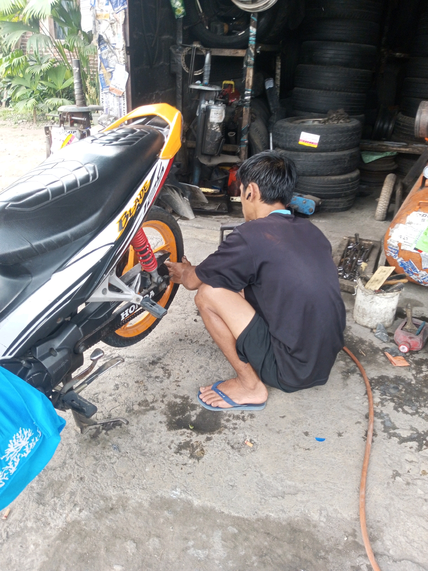 A man is squatting next to a motorcycle, appearing to work on it. The scene is outdoors, with a concrete floor that is wet in places. Several stacks of tires are visible in the background, suggesting the location is a tire shop or repair service. A bright blue tarp is draped over the front of the motorcycle. Tools are scattered on the ground around the man, and a white bucket is also present. The man is wearing a black t-shirt and black shorts, and blue flip-flops. The motorcycle is white and orange, with the word "BLADE" visible on its side. This scene is located in the city of Ngasinan, Indonesia.