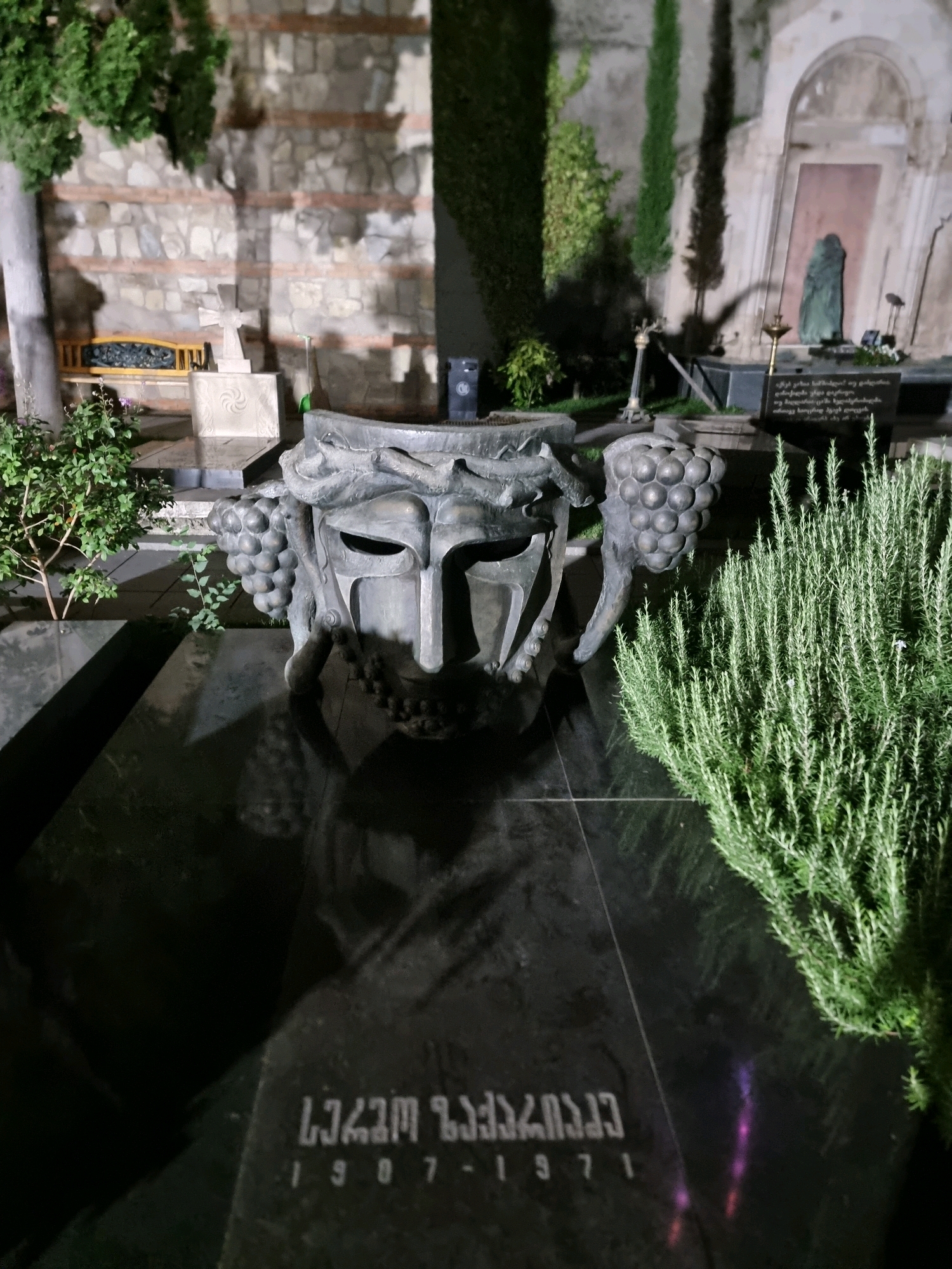 The image shows a close-up view of a grave marker at night. The marker is made of dark, polished stone and features an inscription in Georgian script. Above the inscription is a large, sculptural element that appears to be a mask with grape clusters on either side. In the foreground, to the right, is a bushy green plant, possibly rosemary. In the background, there is a stone wall, a bench, and other grave markers, including a statue of a veiled figure and a small monument with a cross. The lighting suggests it is nighttime, with artificial lights illuminating the scene.