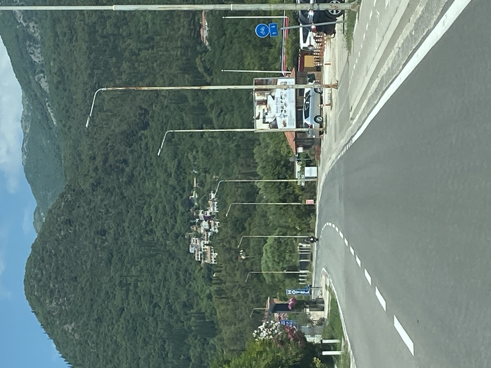 An overcast, daytime, vertical medium shot captures a roadside scene with a winding asphalt road in the foreground, leading towards a hillside densely covered with green trees. A cluster of white-roofed buildings is visible nestled on the upper portion of the treed hillside. The road is marked with dashed white lines. To the left of the road, a sign with the word "HOTEL" is visible, accompanied by some flowering bushes and a concrete structure. A lone motorcyclist is riding on the road in the middle distance. On the right side of the road, several streetlights stand tall, one showing visible rust. A billboard with images and text is positioned behind a small structure with a red roof. A silver car and a darker vehicle are parked near the billboard. Further down the road, a blue sign with bicycle symbols is mounted on a pole. The sky above is a pale blue with scattered clouds, and the background features mountainous terrain.