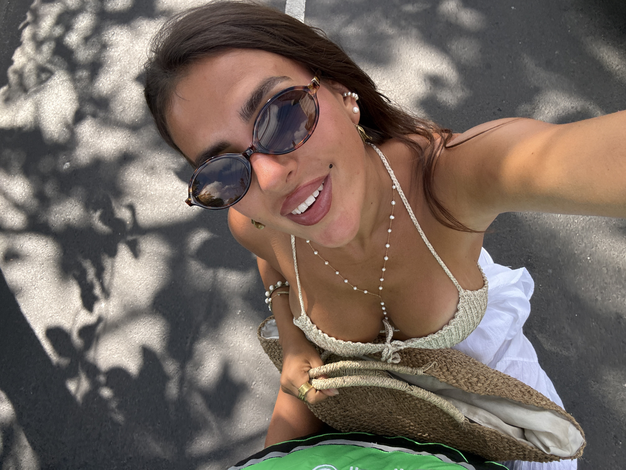 A woman is captured in a high-angle selfie outdoors, her face angled towards the camera. She wears large, tortoiseshell sunglasses and a light-colored, crocheted halter top, adorned with a delicate pearl necklace that drapes across her chest. Her smile is wide, revealing white teeth. A gold ring is visible on her left hand, which holds the strap of a woven straw bag. The bag itself contains a white garment, possibly a dress or skirt. She also has pearl earrings and a small mole below her lower lip. Her hair is dark brown and frames her face. The background consists of a dark, asphalt-like surface, with dappled shadows cast by overhead foliage, suggesting an outdoor setting like a street or pathway in Kuta Selatan, Indonesia.