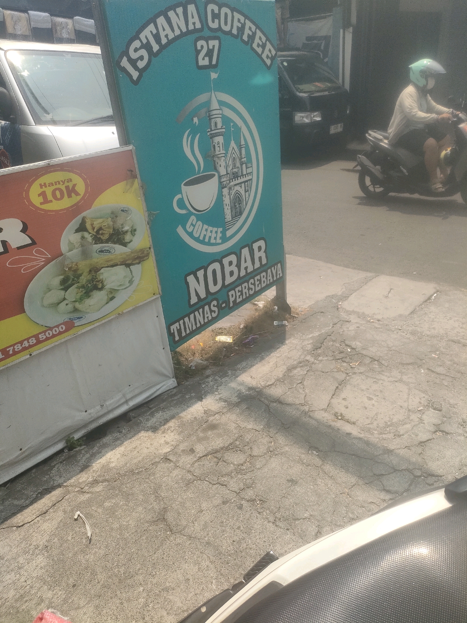 In Surabaya, Indonesia, a turquoise sign for "ISTANA COFFEE 27" stands prominently. The sign features a stylized castle graphic with a coffee cup and steam above the text "COFFEE". Below this, "NOBAR" and "TIMNAS - PERSEBAYA" are visible. To the left, a red and yellow sign advertises "HANYA 10K" (meaning "Only 10K") and displays images of food dishes. A white tarp is situated behind these signs. In the background, a silver car is parked, and a black van is driving on the street. A person wearing a light-colored shirt and a green helmet is riding a motorcycle. The scene is set on a cracked concrete pavement with some dry grass and debris scattered around.