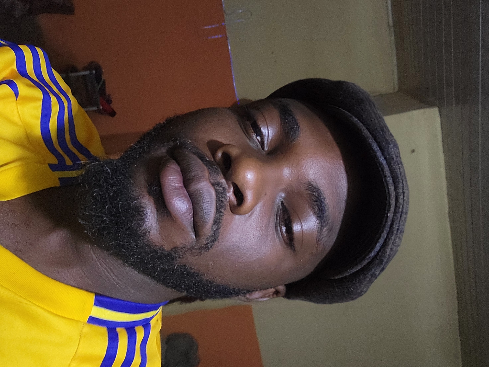 A close-up, waist-up shot features a Black man wearing a dark-colored flat cap and a bright yellow jersey with blue and yellow stripes on the shoulders. He has a dark beard and appears to be indoors. The background is a muted orange wall with some shelving visible. The lighting suggests an indoor setting, possibly at night or in a dimly lit room. There are no other individuals present in the frame. The image is a selfie, likely taken by the subject himself. The subject's facial expression is neutral, and he is looking directly at the camera. The overall environment appears to be a personal living space.