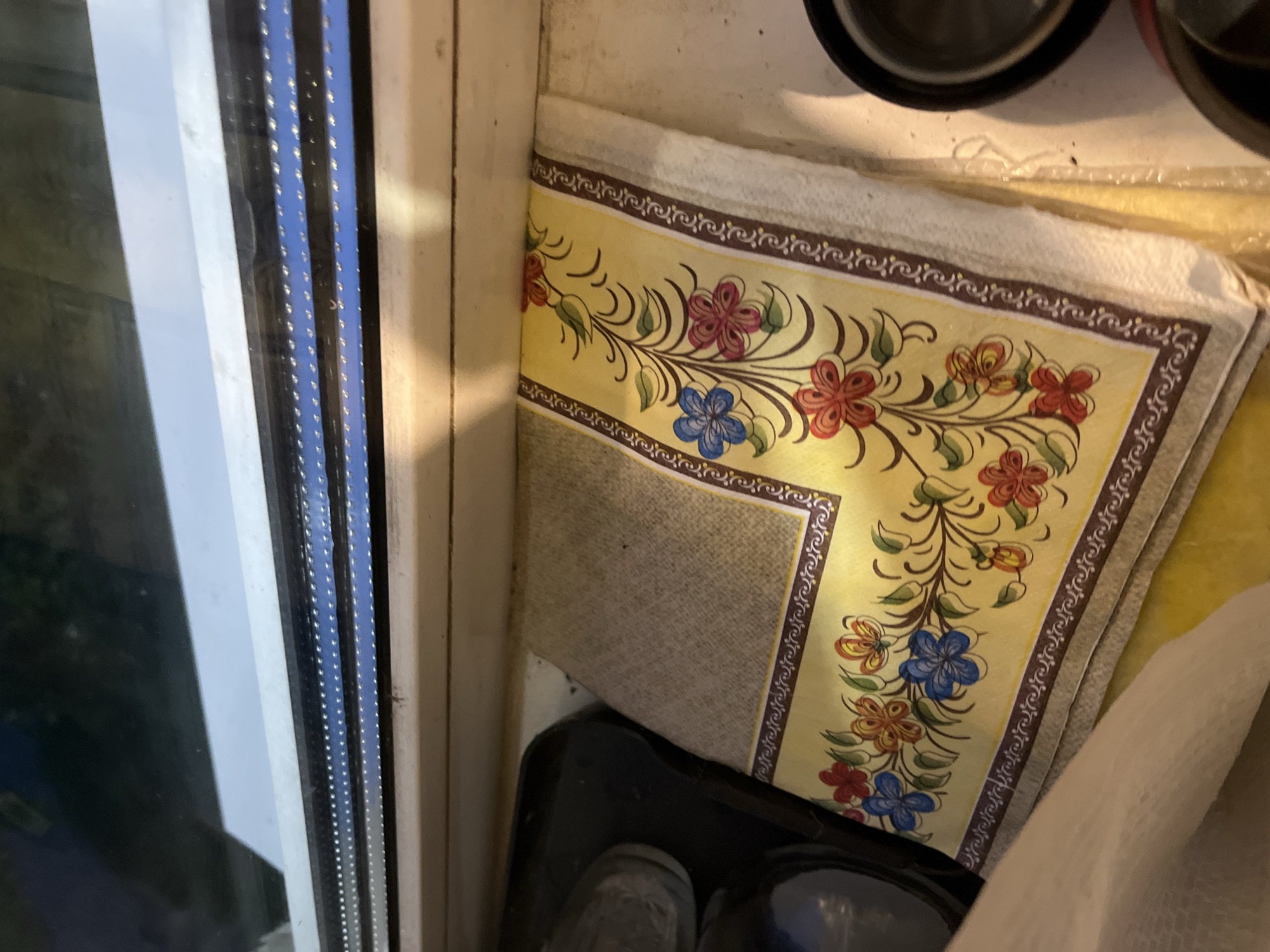 The image depicts a close-up of a decorative cloth, likely a placemat or napkin, with a floral pattern on a yellow background, placed on a windowsill. The cloth is folded and rests on a white surface, possibly the window ledge. To the left, a dark object, possibly a pot or container, is partially visible. The windowsill itself appears to be made of a light-colored material. Above the windowsill, a window is visible, reflecting the sky or an interior light. The scene is captured indoors, suggesting a domestic or commercial setting. The overall impression is one of everyday objects within a functional space, with the decorative cloth adding a touch of visual interest. There are no people or animals present in the image. The lighting suggests it may be daytime or an area with artificial light. There are no visible indications of the specific location within Minsk, Belarus, other than the implied setting of a windowsill.