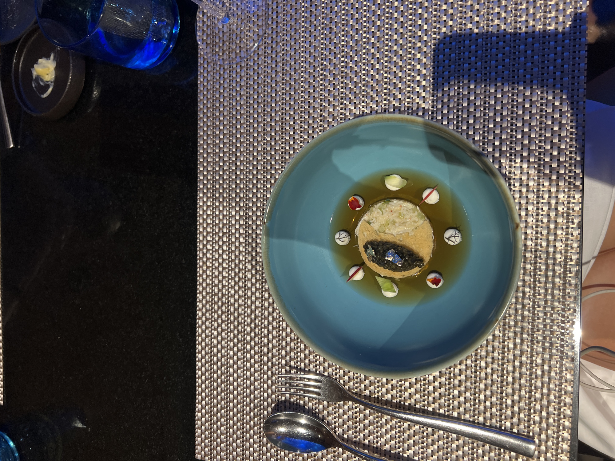 A restaurant table in Kuta Selatan, Indonesia, shows a blue plate containing a round food item topped with caviar and surrounded by small white, red, and green garnishes in a shallow sauce. Cutlery is placed below the plate on a woven placemat. The table is also set with blue glassware and a small dish with a white condiment. The adjacent table is black, and a person's arm wearing white is visible on the right side.