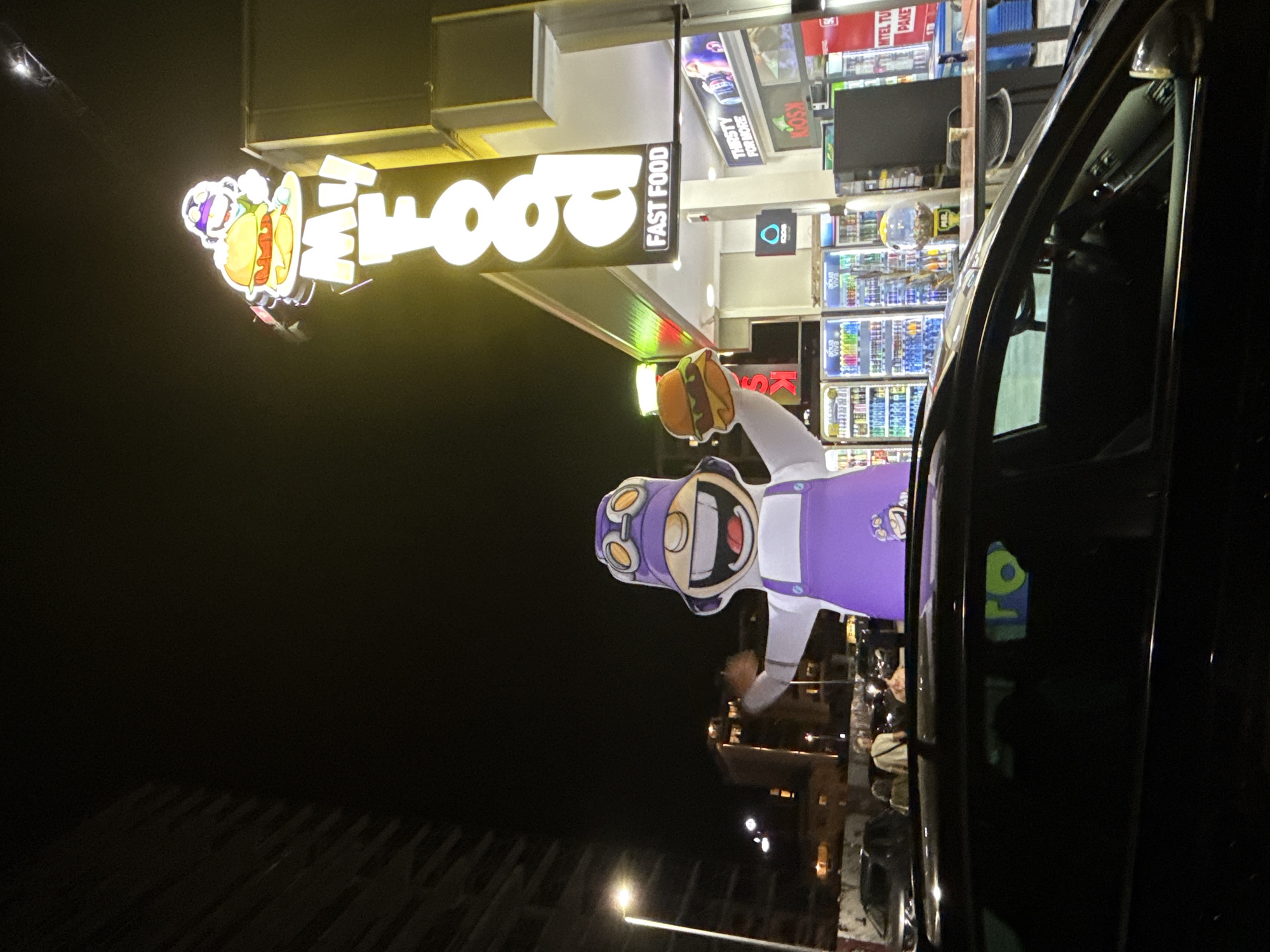 An inflatable mascot holding a burger stands in front of a fast-food establishment at night in Budva, Montenegro. The mascot, wearing a purple outfit and goggles, is positioned in the foreground. To the right, a neon sign displays "My Food" with the words "FAST FOOD" beneath it. Behind the mascot, refrigerated display cases filled with beverages are visible, along with a kiosk. The scene is illuminated by streetlights and the neon signage. A dark-colored car is parked in the foreground, partially obscuring the lower part of the inflatable mascot. In the background, building lights and streetlights indicate an urban setting.