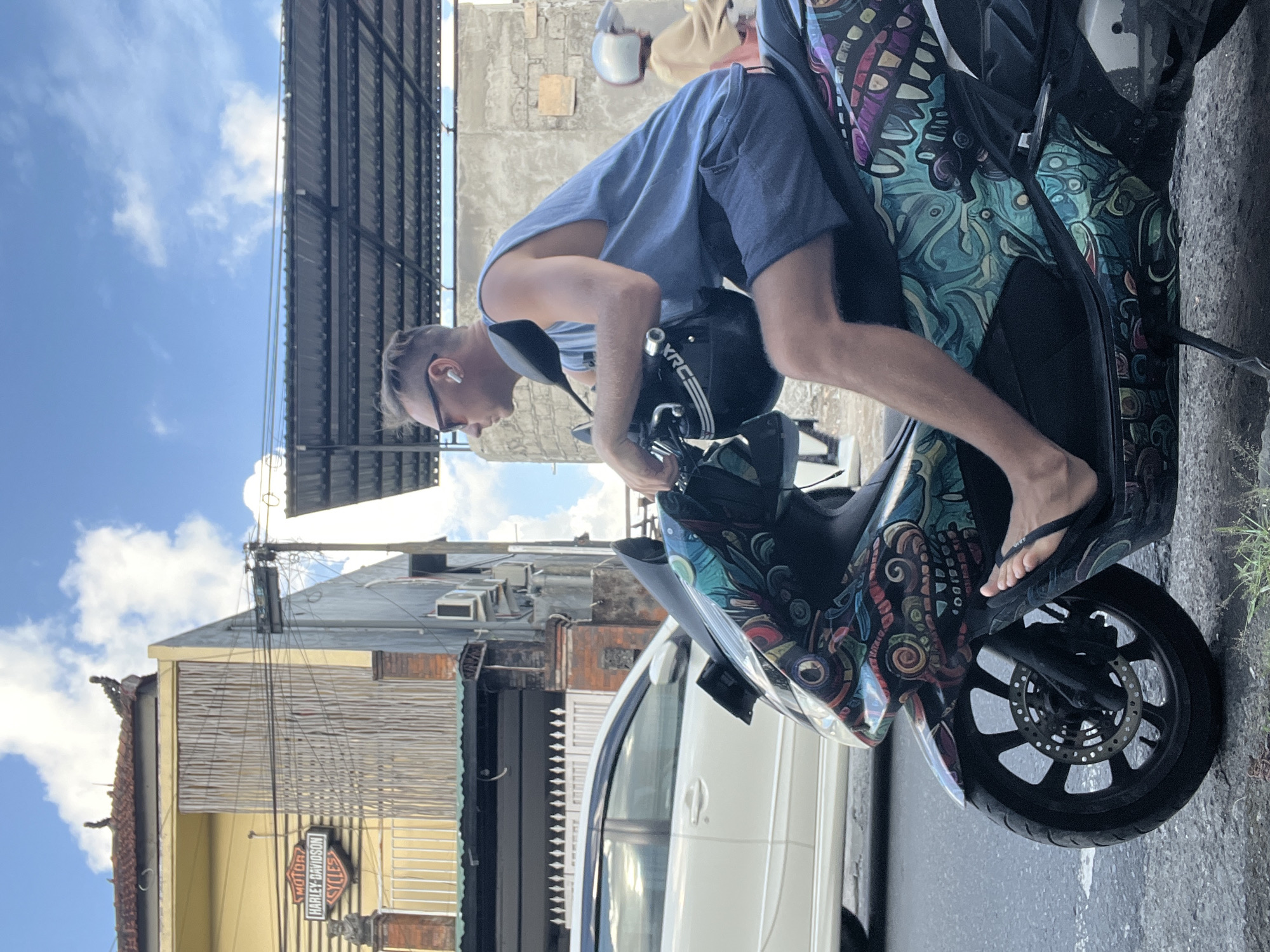 A vertical shot shows a man inspecting a parked motorcycle in the Ubud District of Indonesia.
The man, wearing a blue tank top and blue shorts, leans over the motorcycle's handlebars. He wears sunglasses and earbuds. His right foot is visible on the footrest, clad in a black flip-flop. The motorcycle has a colorful, patterned design.
Behind the motorcycle, a white car is partially visible. The background includes buildings with various architectural styles, typical of urban areas in Ubud. A Harley-Davidson sign is visible on one building. Overhead cables and infrastructure are seen against a blue sky with scattered clouds.