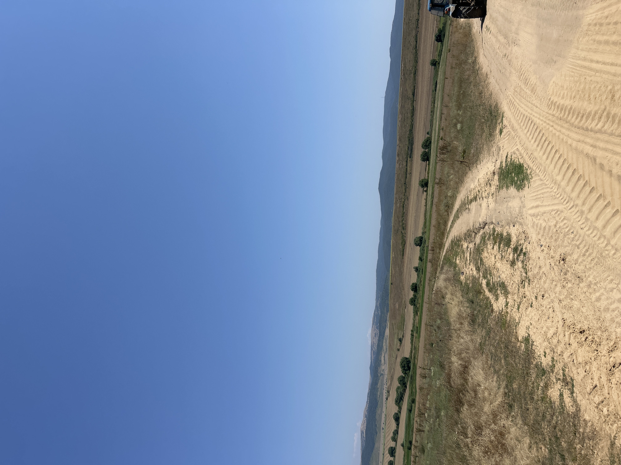 The image captures a vast, rural landscape under a clear, bright blue sky. A dirt road, marked with distinct tire tracks, dominates the foreground, winding its way through dry, golden grass and patches of greener vegetation. To the left of the road, the terrain slopes gently upward, dotted with scattered trees and fields that appear to have been recently harvested.
In the middle ground, a series of rolling hills recede into the distance. These hills are a mix of brown and green hues, suggesting agricultural land or natural vegetation. The horizon is marked by a range of larger, darker hills, some of which have a rugged, almost mountainous appearance. A thin line of white or light-colored clouds is visible along the crest of one of the distant hills.
On the right side of the frame, a blue tractor is partially visible, its rear wheel and part of its chassis facing the camera. The tractor appears to be stationary on the dirt road. The overall impression is one of an open, agricultural expanse, possibly a farm or rural area on the outskirts of a settlement. The clear sky and dry vegetation suggest a warm, sunny day, likely during the summer months.
This scene is consistent with the agricultural landscape found in and around the village of Poroy, Bulgaria, which is known for its rural setting and farming activities.