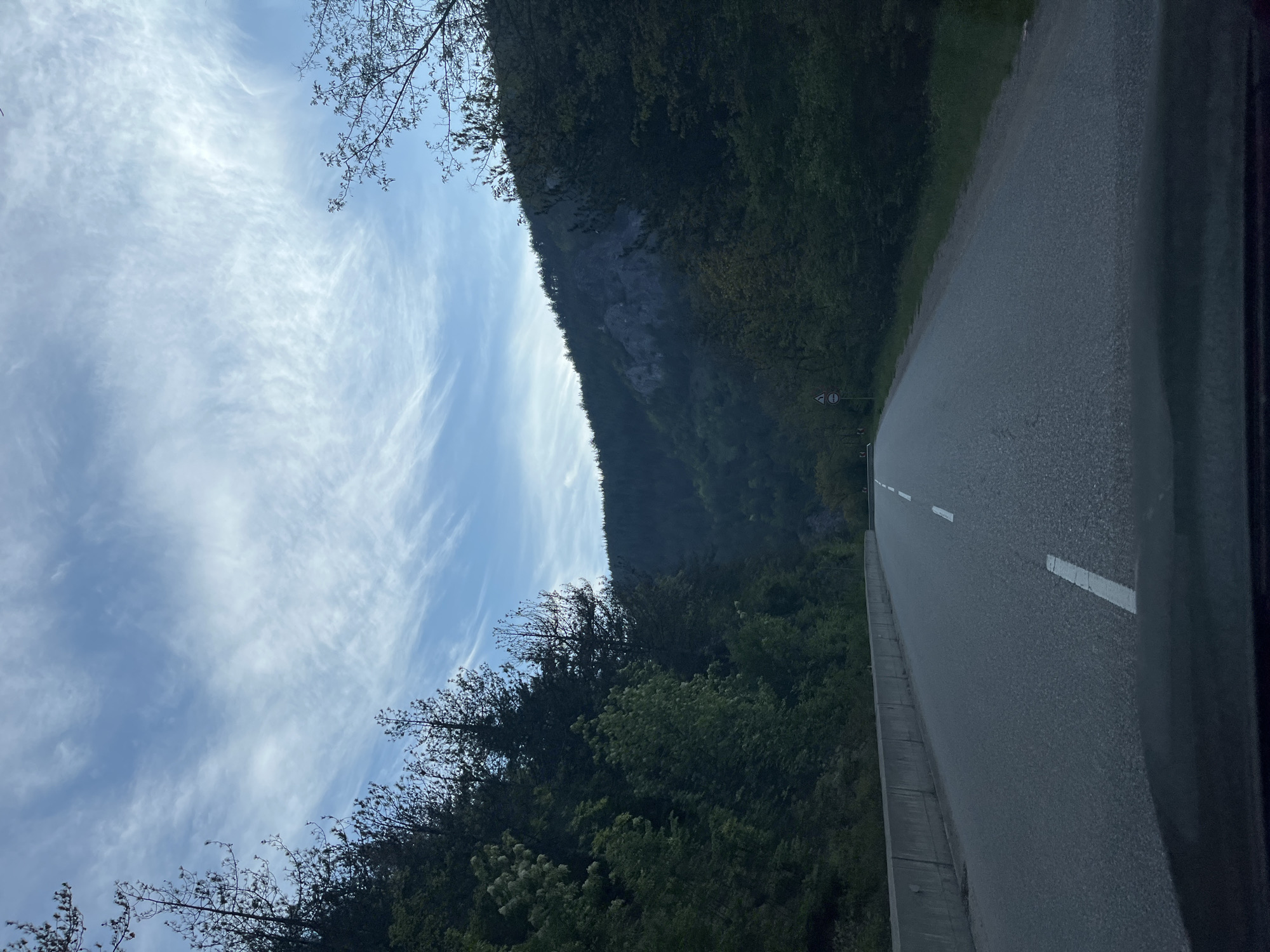The image shows a road in a mountainous, forested area. To the right of the road, there is a dark green forest. To the left of the road, there is a mountain. Above the mountain, there is a blue sky with white clouds. There are no people visible in the image. The scene may be in Hvoina, Bulgaria.