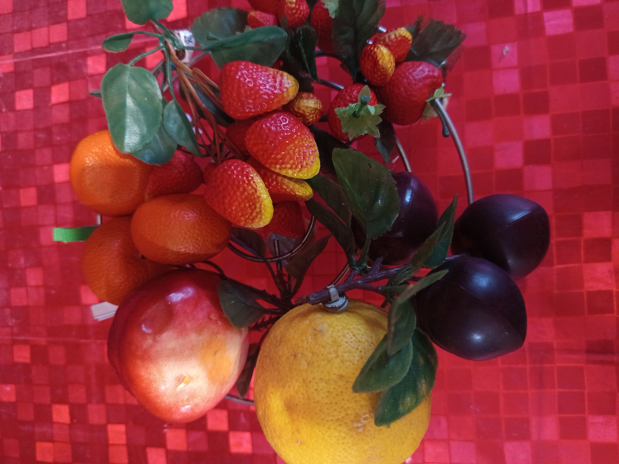 A still life arrangement of artificial fruits is displayed on a red checkered surface. The fruits include several bright red strawberries with yellow tips, a cluster of small orange tangerines, dark purple plums, a red and yellow apple, and a yellow lemon. The fruits are attached to a wire frame with artificial green leaves and brown branches. A small white label with the number "26" is visible on one of the branches. The lighting suggests an indoor setting, and there are no discernible weather or time of day cues. The overall impression is one of a decorative display rather than a real fruit selection. The location context provided is Pavlohrad, Ukraine.
