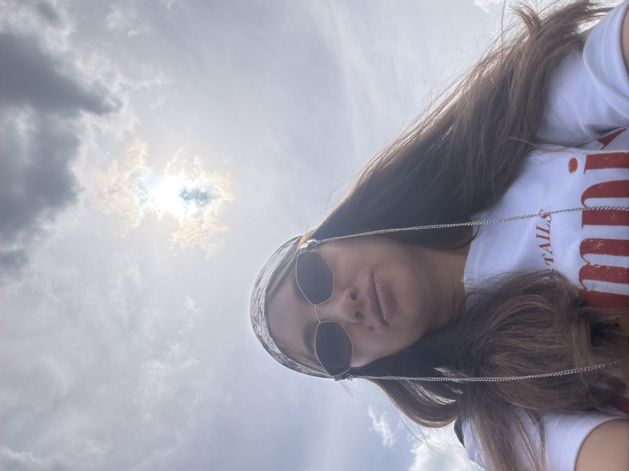 A low-angle, waist-up shot captures a person with long, dark hair wearing a white t-shirt and a patterned headband. The person is also wearing angular sunglasses with decorative chains. The sun is partially obscured by clouds in the background, creating a bright, hazy sky. The t-shirt has red lettering, with "TAILS" partially visible above a larger word that appears to be "MMUNI". The image was taken outdoors, possibly in or near Kremenkul', Russia, given the cloudy sky typical of the region.