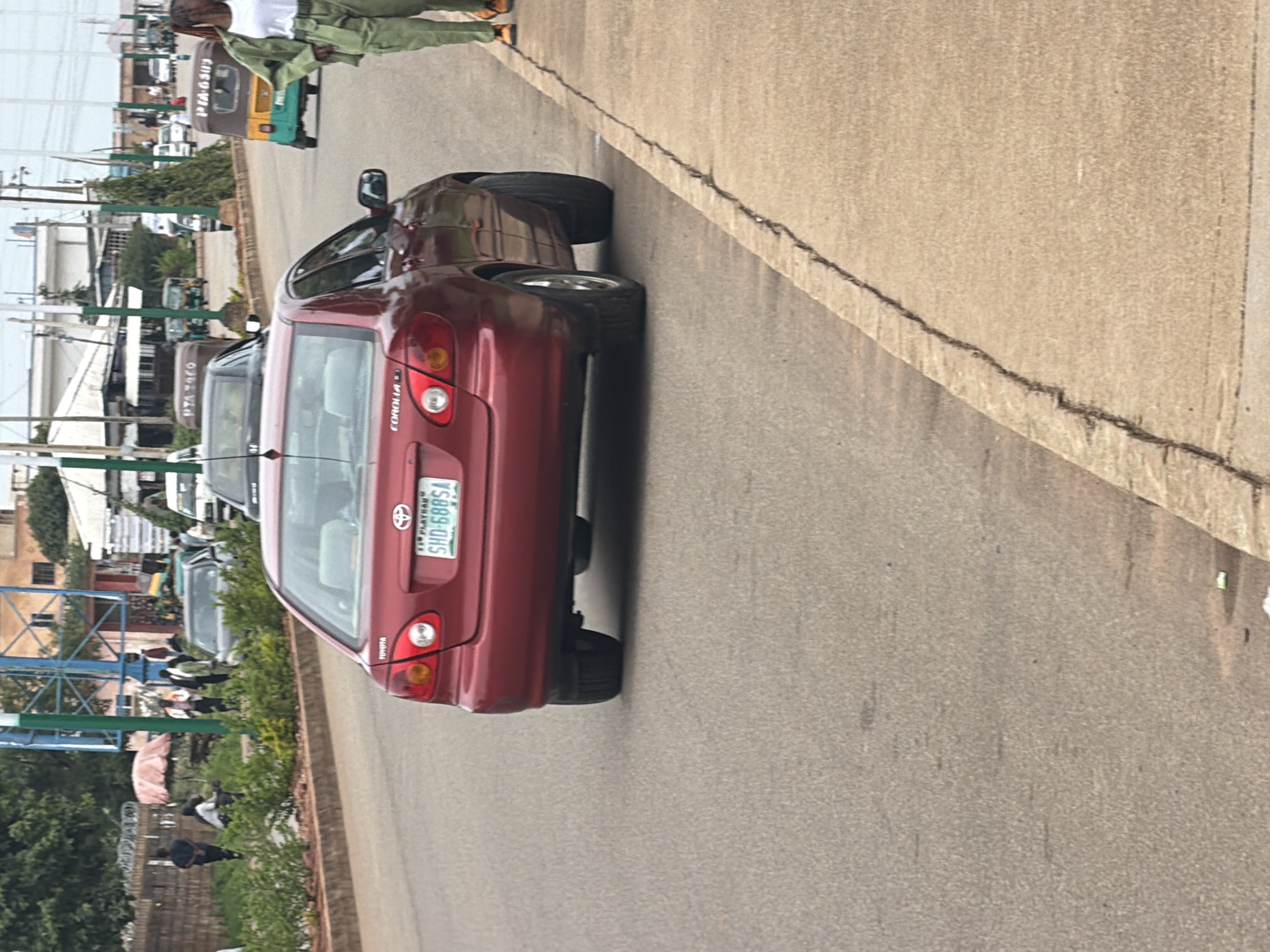 This is a vertical, eye-level shot of a street scene in Bukuru, Nigeria. A dark red Toyota Corolla is parked on the right side of the frame, facing away from the camera. The car's license plate, visible on the rear, reads "SHD-6885A" and appears to be from Plateau State. To the left of the car, a paved road curves away from the viewer. Beyond the road, a sidewalk area is lined with lush green bushes and trees. Several people are visible walking and standing in this area. In the background, buildings and other vehicles, including what appears to be a three-wheeled auto-rickshaw, suggest an urban environment. The lighting indicates it's daytime, likely late afternoon judging by the light quality. The overall scene depicts a common street view in a Nigerian town, with everyday activity.