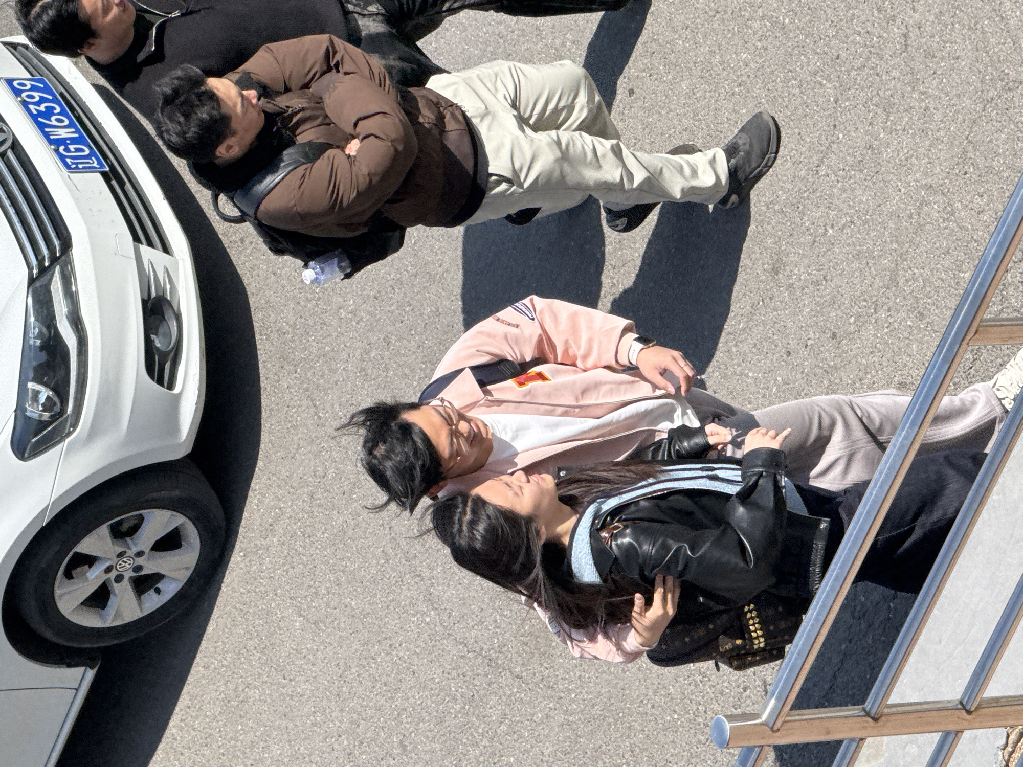 The image shows an overhead view of a street scene. A white car is parked on the left, displaying the license plate "辽G·W6399". Three individuals are lying on the pavement. One man, wearing a brown puffer jacket and beige pants, lies above another man in a black shirt. Below them is a couple; the man wears a pink jacket, and the woman wears a black leather jacket. Part of a metal railing is visible on the right. The location is Zhongtun, China.