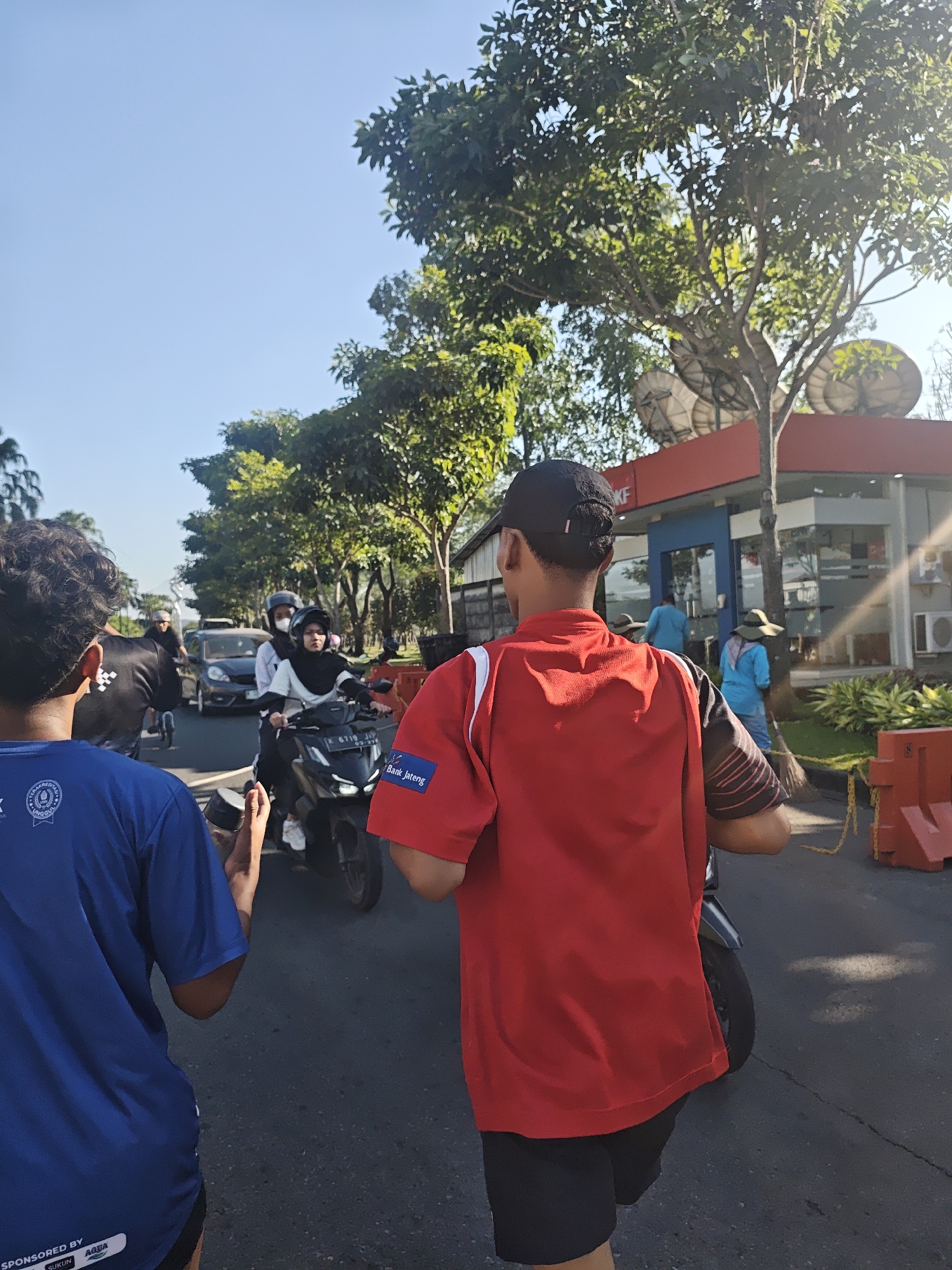 In Bacin, Indonesia, two young men are walking on a road, likely participating in or observing an event. The man in the foreground, on the right, is wearing a red t-shirt with black and white striped sleeves and black shorts. He has a black baseball cap on his head, and his back is towards the camera. The man on the left is wearing a blue t-shirt and black shorts. In the background, several other individuals are present, including people riding motorcycles. One woman on a motorcycle is wearing a hijab and a face mask. Traffic is visible, with cars and other motorcycles on the road. Trees line the street, and a building with a red awning and satellite dishes is visible in the distance. The sky is clear and blue, indicating it is daytime.