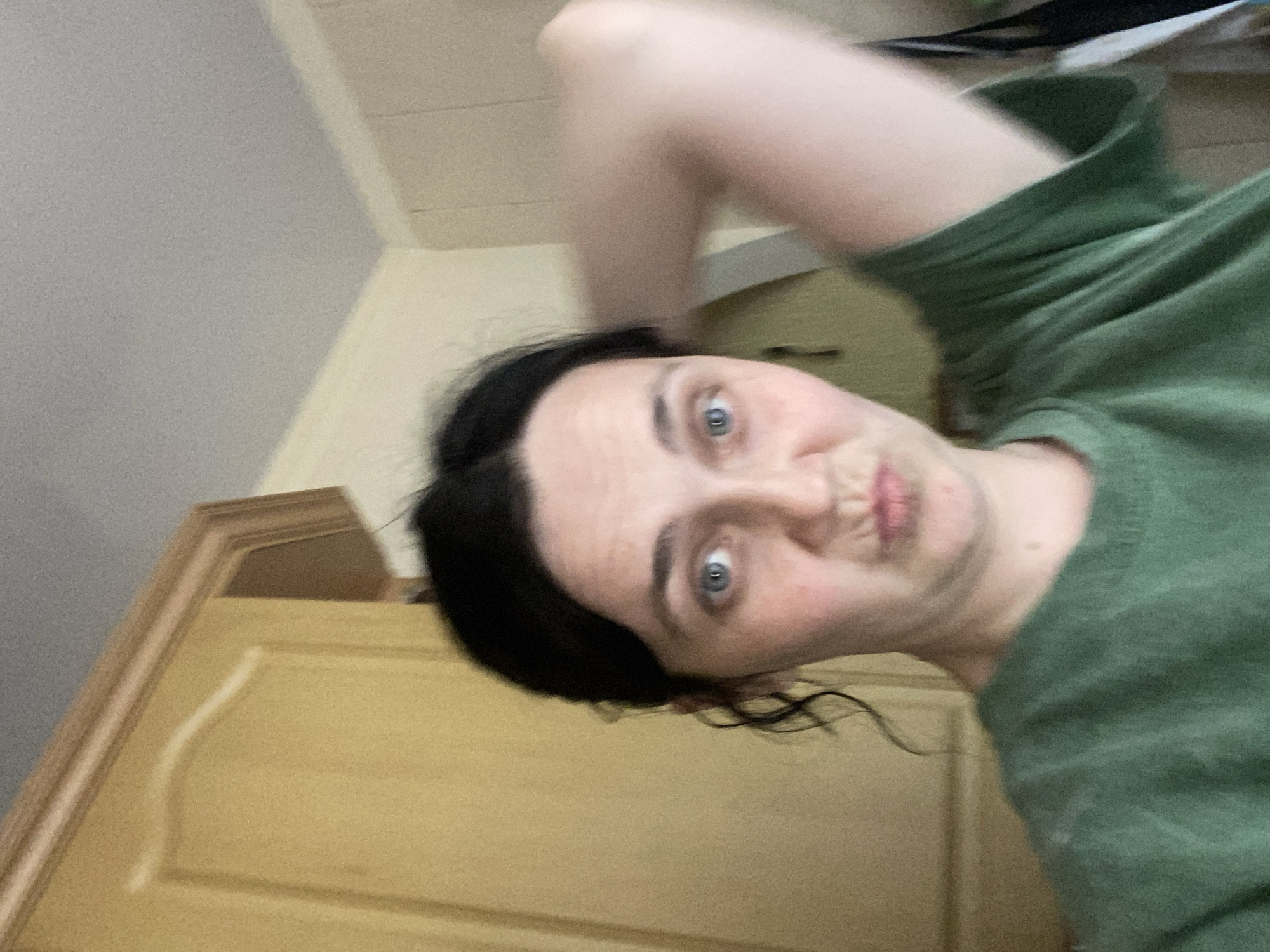 The media file is an image of a young woman with dark hair. She is wearing a green long-sleeved shirt. Her left arm is extended upwards, and she is holding her phone to take a selfie. Her head is tilted, and she is making a pouting expression. Her eyes are open and looking towards the camera. The background is blurred but appears to be indoors, with a closet door and a wall visible. The lighting suggests it is daytime, but there are no specific cues to indicate the time of day or weather. The woman's expression conveys a playful or humorous emotion. There is no visible text in the image.