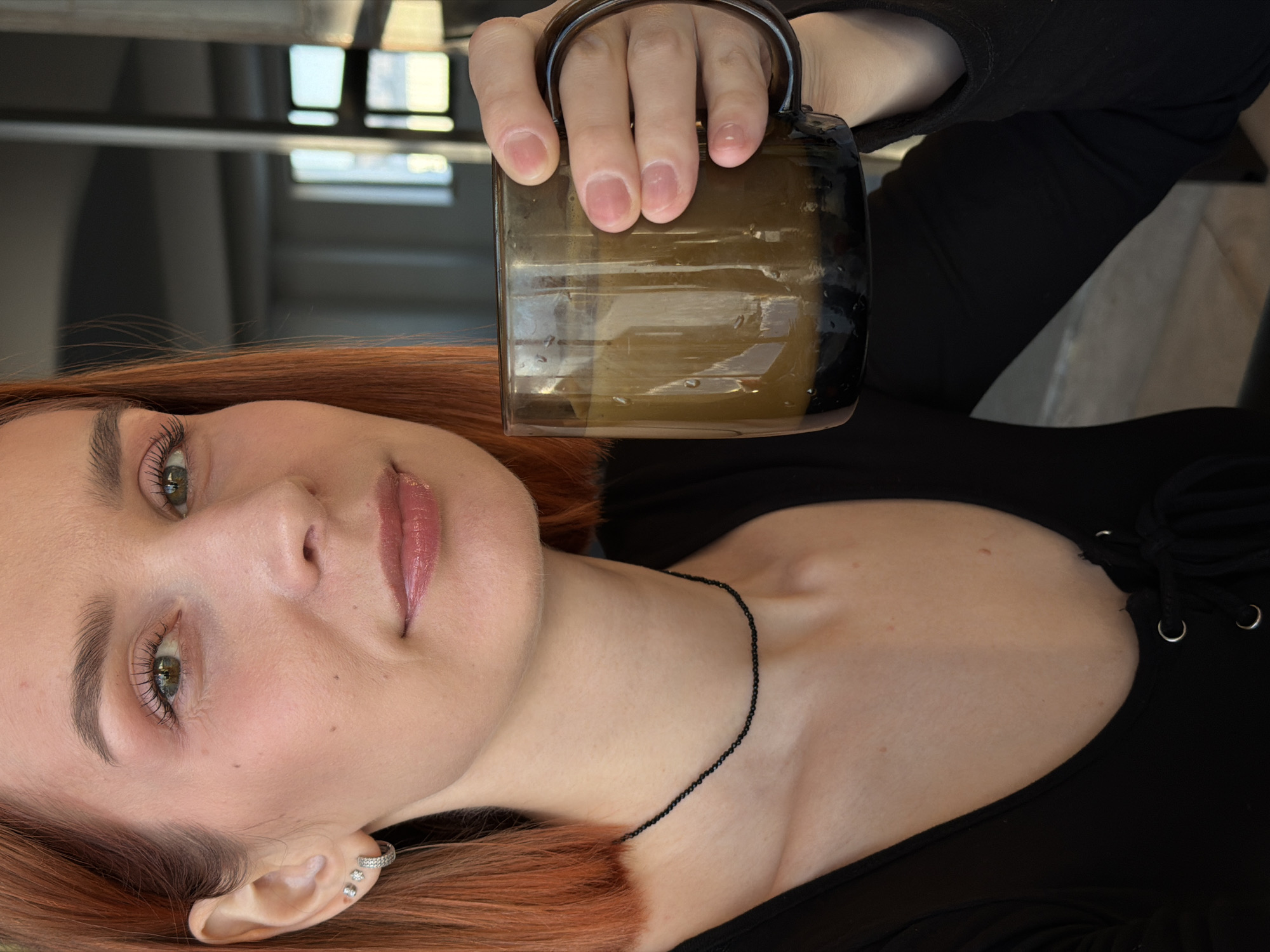 A young woman with light skin, red hair, and green eyes is holding a brown glass mug with a handle. She is wearing a black, low-cut, long-sleeved shirt. A thin black necklace is visible around her neck, and she has a small earring in her left ear. The background includes what appears to be part of a stairwell, with a window visible in the distance. There is no information available in this image to indicate the location is Chaikovsky, Russia.