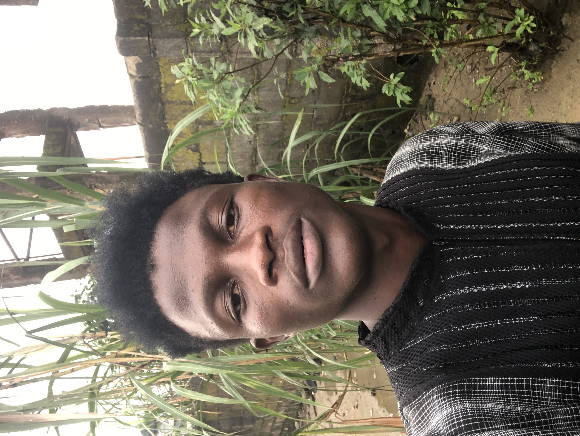 In Ado, Nigeria, a young Black male takes a close-up selfie outdoors. He has dark skin, short black curly hair, and is wearing a black garment with a white checkered and striped pattern. The background shows tall green reeds on the left, and on the right, a rough, weathered brick wall with some green foliage growing against it. The ground appears to be dirt and possibly wet. The sky is overcast and bright. The overall impression is of a candid shot taken in a somewhat rustic or natural setting within the city.