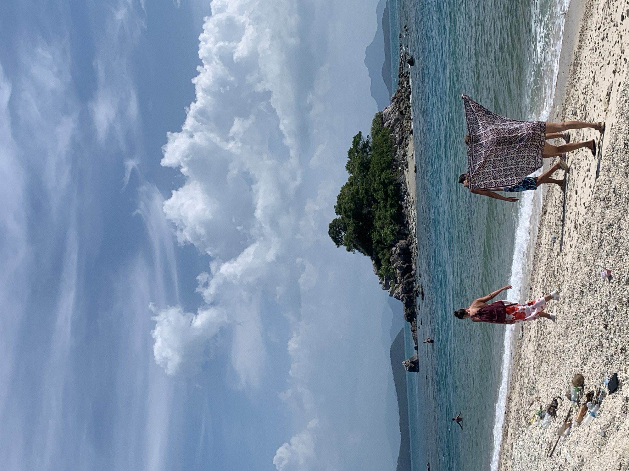 A group of people are on a beach in Thailand. In the foreground, two people are walking towards the water, one of whom is holding up a patterned cloth. Another person, wearing a red and white floral skirt, walks away from the water. The beach is littered with plastic bottles and other debris. In the background, a small, rocky island with trees is visible in the blue ocean. Distant islands and mountains are seen on the horizon under a sky with white clouds.