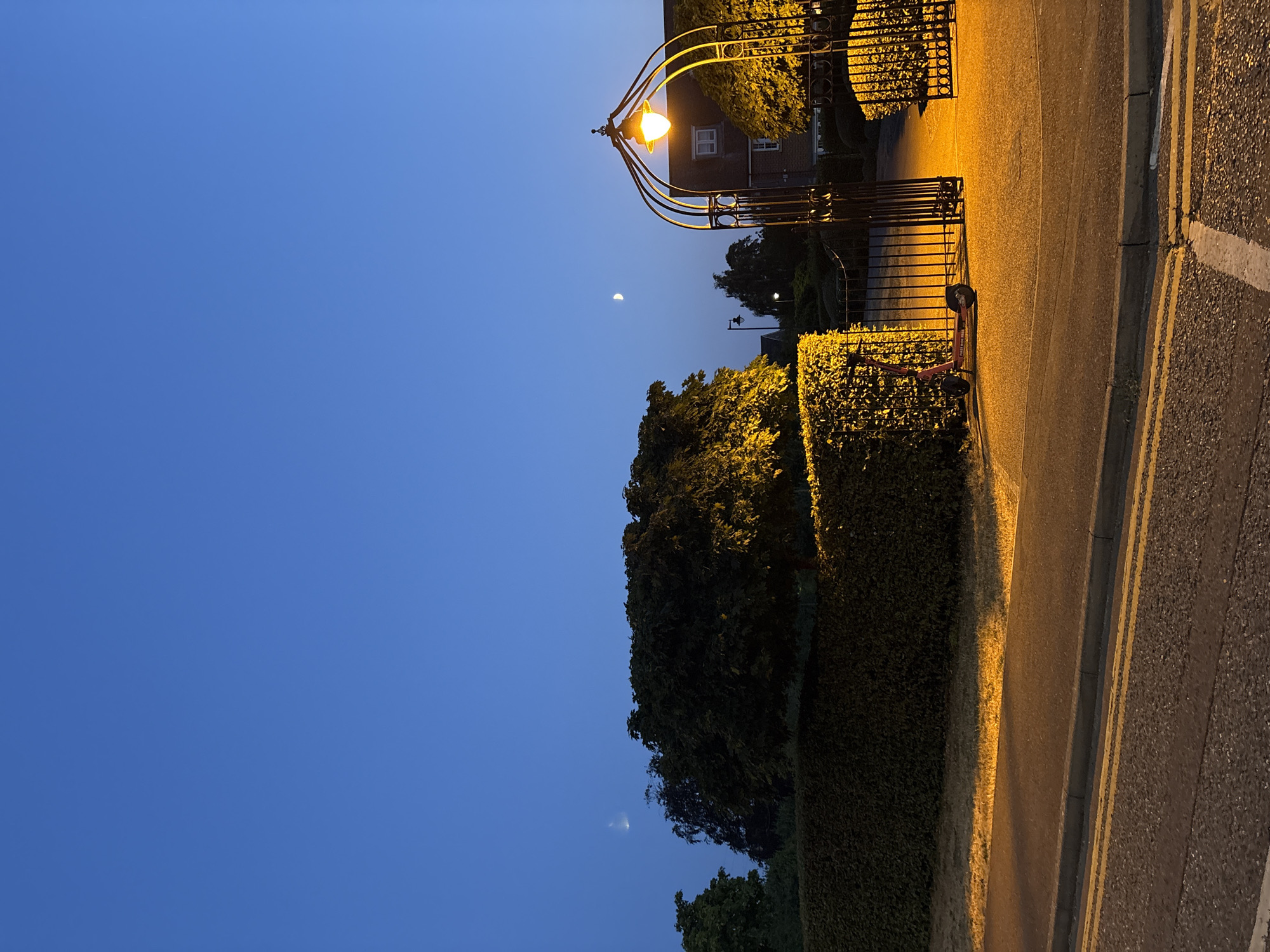 The image shows a street scene at dusk in Chelmsford, United Kingdom. A streetlamp illuminates an ornate wrought-iron gate, which is open. To the left of the gate, a red electric scooter is parked on a paved surface. Behind the gate and scooter, a well-maintained hedge forms a backdrop. A tree with illuminated foliage stands to the left of the gate. In the background, a building with a pitched roof is visible. The sky is a deep blue, with two crescent moons faintly visible. The street in the foreground has double yellow lines and a curb.