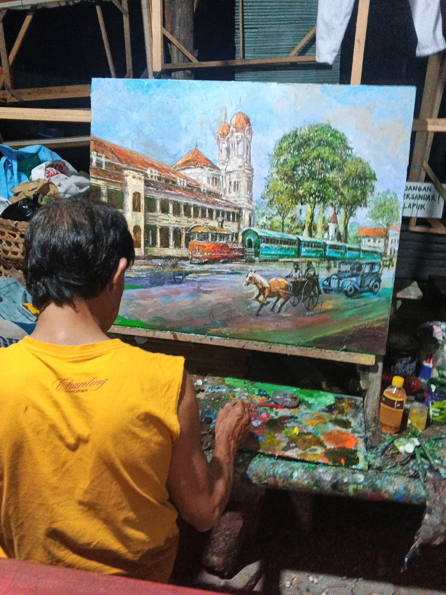 The image depicts a man painting a vibrant oil on canvas artwork of a historical street scene, likely located in Ungaran, Indonesia, given the distinct architectural style and the presence of colonial-era elements. The painting showcases a grand building with a domed tower, an orange tram, a green tram, a horse-drawn carriage, and an early automobile, set against a backdrop of lush trees and a bright blue sky. The artist, wearing a yellow sleeveless shirt, is actively engaged in his craft, his palette laden with various colors. The setting appears to be an artist's studio or workspace, with the painting propped on an easel and art supplies scattered around. The background includes wooden structures and what looks like corrugated metal sheeting, suggesting a rustic or utilitarian environment. A sign with Indonesian text, "JANGAN ERSANDAR LAPUK," is visible on the right, further hinting at the local context. The overall impression is one of focused artistic creation within a lived-in, creative space.