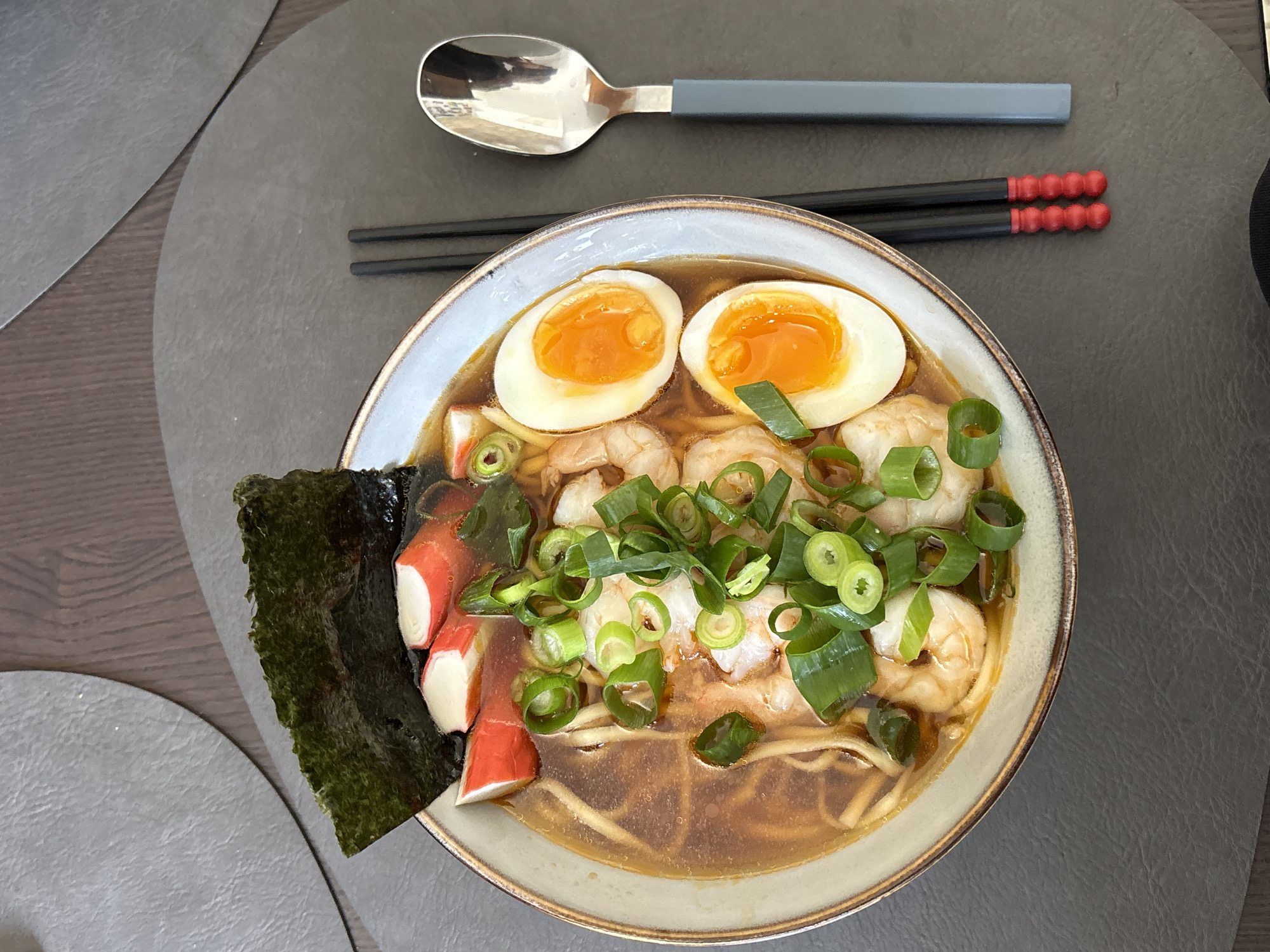 A top-down, medium shot shows a bowl of ramen on a grey placemat placed on a brown wooden table. To the left of the bowl is a dark green piece of seaweed and three pieces of what appear to be crab meat. The bowl itself contains ramen noodles, shrimp, sliced scallions, and two halved eggs with orange yolks. Above the bowl, a silver spoon with a grey handle and a pair of black chopsticks with red ends are visible. A second grey placemat is partially visible on the left side of the frame.
The photograph was likely taken indoors. The location within Ramat Gan, Israel cannot be determined from the image.