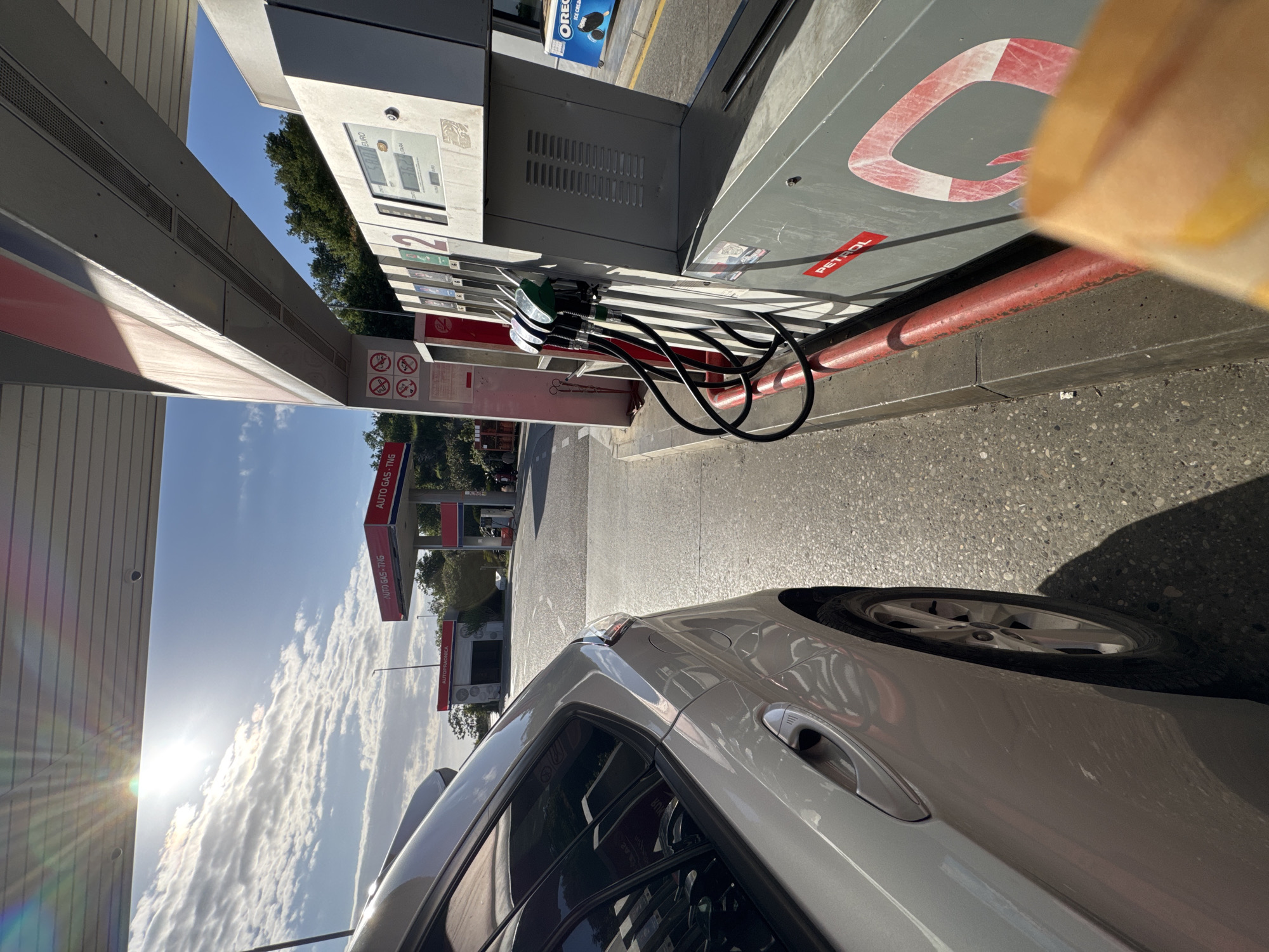 The image shows a car parked at a petrol station in Petrovac na Moru, Montenegro. The car is light grey, and the fuel pump is visible, bearing the logo of the Petrol brand, and labels “AUTO GAS-TNG”. In the background are the canopies of other petrol pumps and the service station building. A sign advertising Oreo ice cream is affixed to the side of the station building. The sky is blue with some clouds.