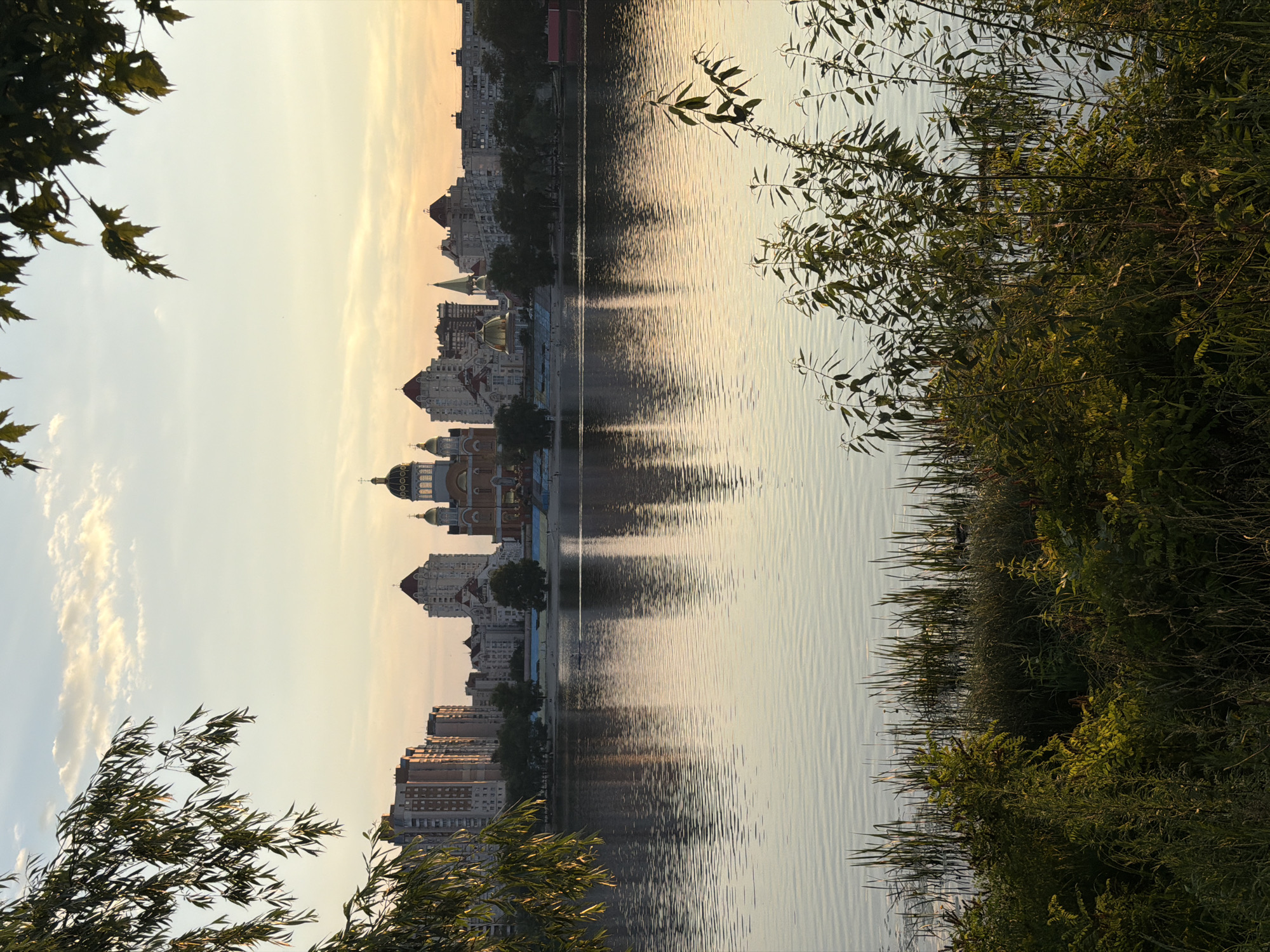 This is a scenic view of the Dnipro River in Kyiv, Ukraine, taken at sunset. The foreground is dominated by lush green vegetation and reeds along the riverbank. The calm water reflects the sky, trees, and the city skyline. In the mid-ground, the cityscape features a prominent Orthodox church with golden domes, surrounded by modern residential buildings. The sky is a soft blend of yellow, orange, and pale blue, with scattered clouds. The scene evokes a peaceful and serene atmosphere.