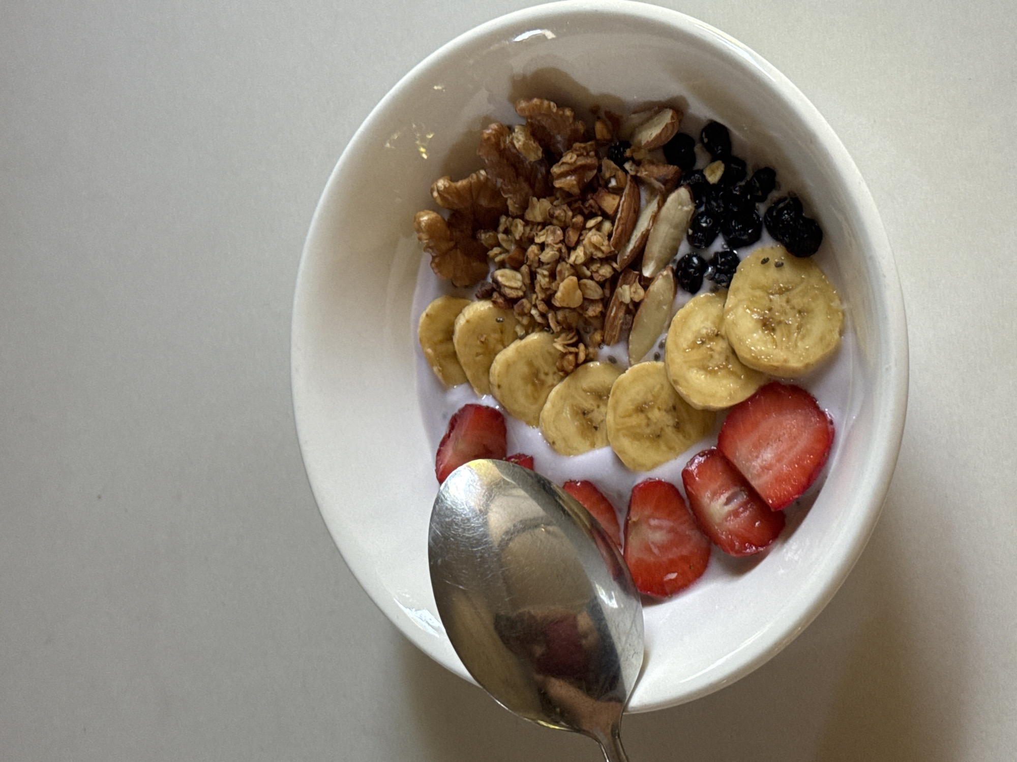 A close-up shot shows a white bowl filled with a healthy breakfast of yogurt, sliced bananas, strawberries, blueberries, granola, and nuts. A silver spoon rests on the left side of the bowl, partially submerged in the yogurt. The background is a plain, light-colored surface, likely a table. The food is arranged artfully, with the fruits forming a colorful crescent on one side and the granola and nuts clustered on the other. The yogurt appears creamy and smooth, with a slightly purple hue, suggesting it might be berry-flavored. The overall impression is one of a fresh, nutritious, and appealing meal. The image does not provide any specific details about its location in Davao City, Philippines, as the background is generic and there are no identifying landmarks or elements.