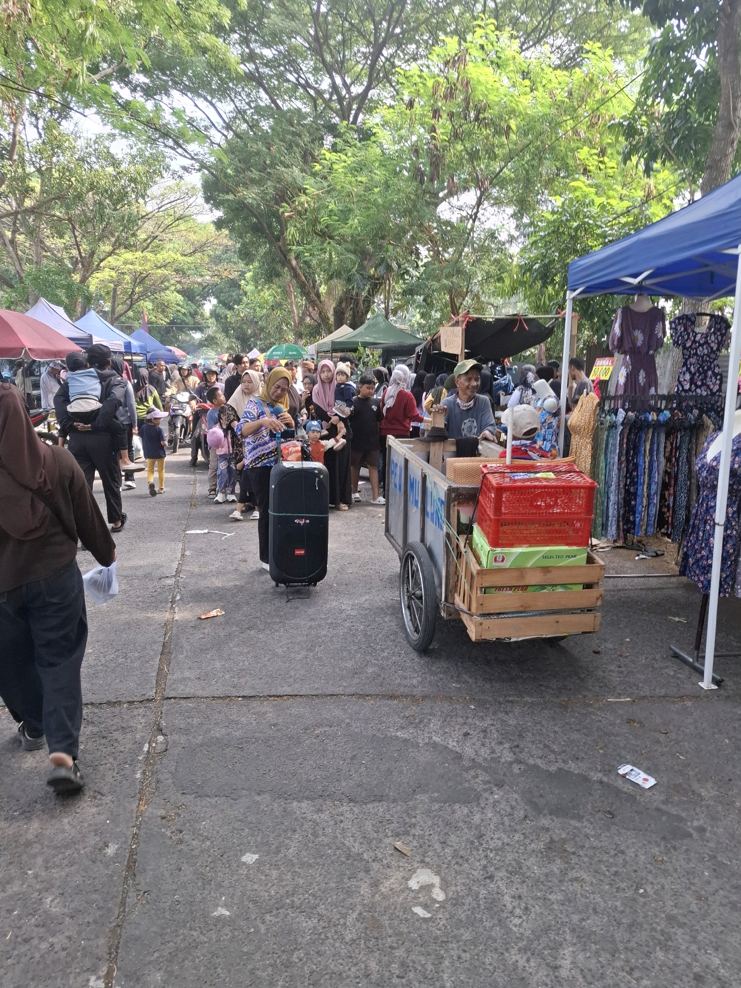 The image depicts an outdoor market scene in the city of Karangtengah, Indonesia, under a canopy of trees. In the foreground, a person wearing a brown headscarf and dark pants walks away from the camera on a paved surface, holding a white plastic bag. To the right of the pedestrian, a large black speaker stands on a metal tripod. Further into the market, a man with a green cap is seated at a cart laden with goods, including red crates and boxes of what appear to be fruit. Behind him, several stalls are set up, displaying clothing for sale. A crowd of people, many wearing headscarves, are browsing and walking through the market. In the background, motorbikes are visible, suggesting transportation options for shoppers. The environment is characterized by a mix of natural foliage and man-made market structures.