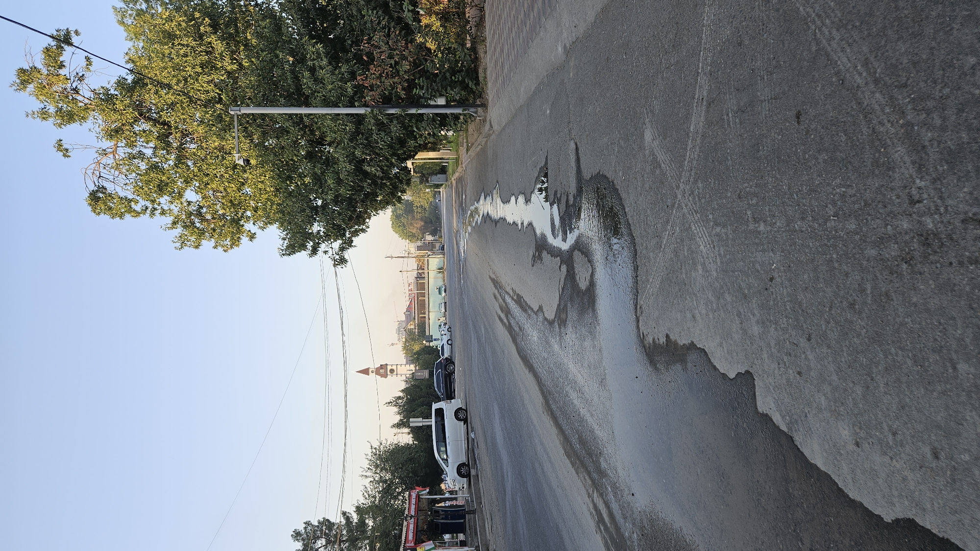 The image depicts a street scene in Dushanbe, Tajikistan, likely in the morning or late afternoon due to the soft lighting. A wet patch of water is prominent on the asphalt road, with some debris, possibly leaves, visible within it. The road shows tire marks. On the left side of the road, there are several parked vehicles, including a white minivan and a black sedan. A small kiosk or shop with signage is also present. Across the street, a clock tower with a red roof is visible in the distance, a distinctive landmark. Lush green trees line the right side of the street, and a lamppost stands tall next to them. A tiled sidewalk runs along the right edge of the road. The sky is clear and blue, suggesting fair weather. The overall impression is of an urban environment during a calm period.
