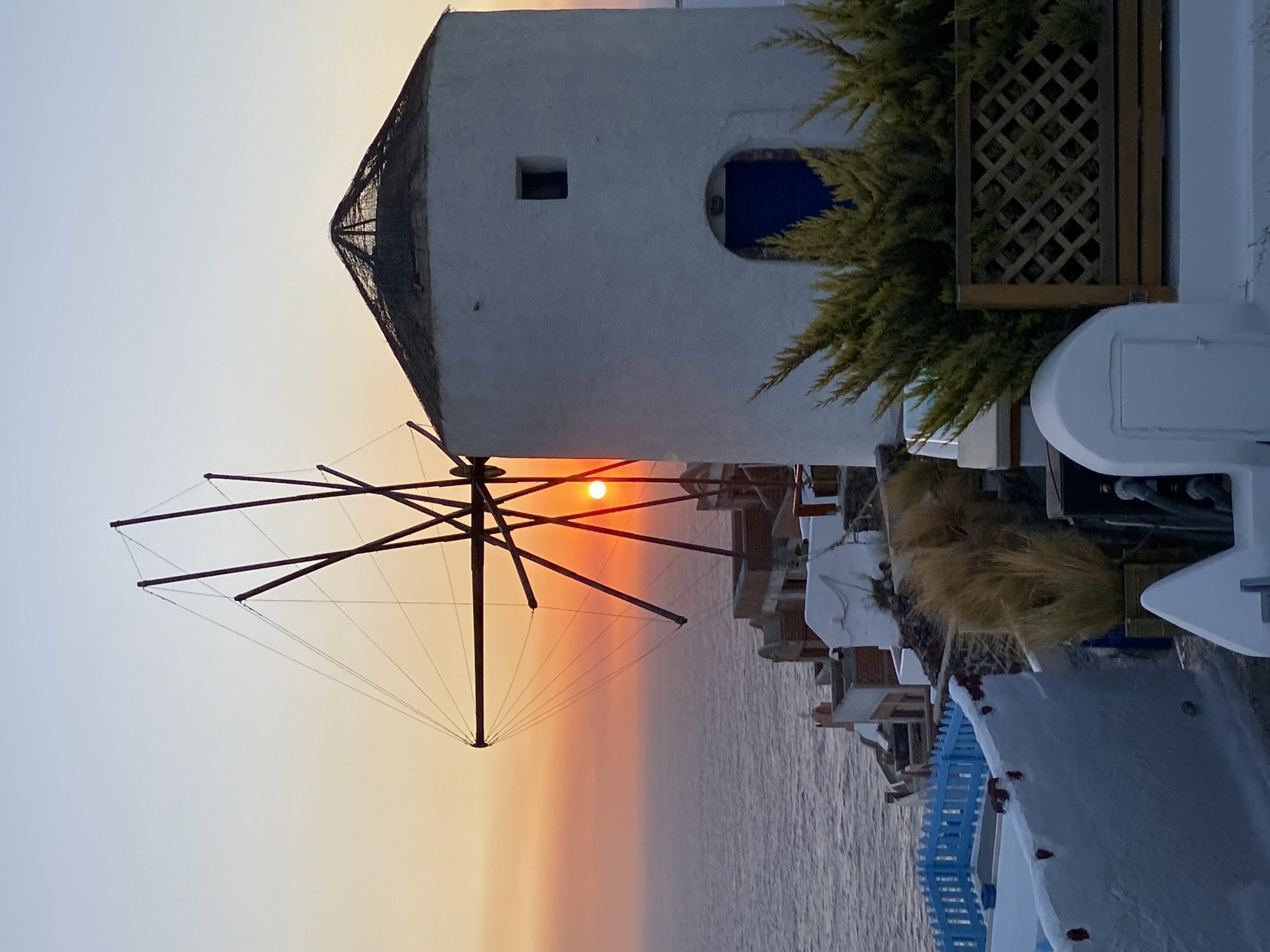 The image depicts a sunset scene in Oia, Municipal Unit of Ia, Greece. A white windmill with dark wooden sails stands prominently in the foreground, partially silhouetted against the vibrant orange and yellow hues of the setting sun. The sun itself is visible as a bright, red disc peeking through the windmill's sails. To the right of the windmill is a white, cylindrical building with a conical roof and a blue door. The structures are situated on a cliff overlooking the Aegean Sea, which appears calm and reflects the colors of the sunset. In the lower portion of the image, there are white buildings with blue railings and some greenery. The overall scene conveys a peaceful and picturesque view of a Greek island village at dusk.