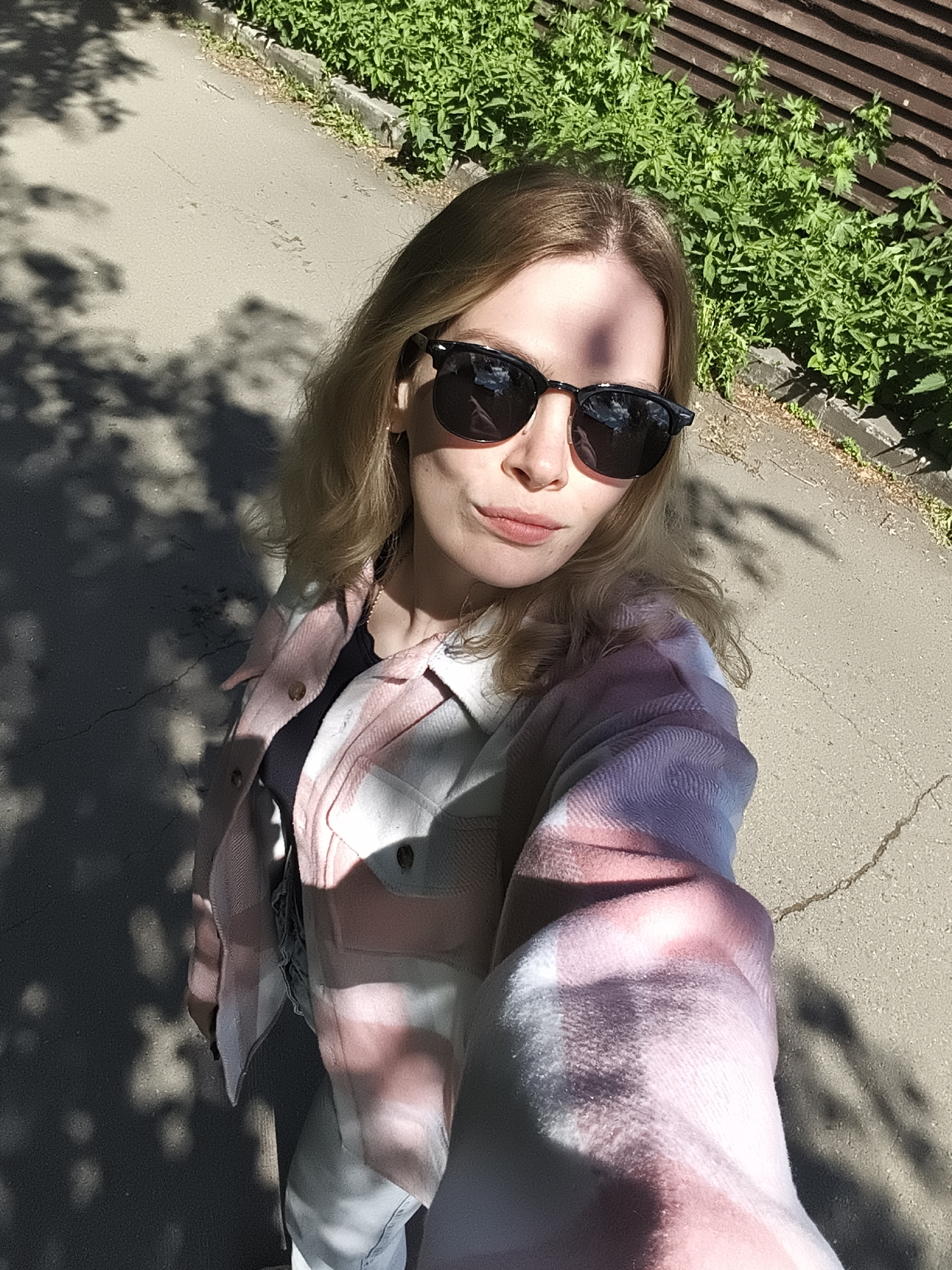 A woman in sunglasses and a pink and white plaid jacket is taking a selfie outdoors on a paved area. The background includes greenery, a wooden fence, and a cracked pavement surface, which suggests a street or park environment. The exact location in Izhevsk, Russia cannot be identified based on the available information.