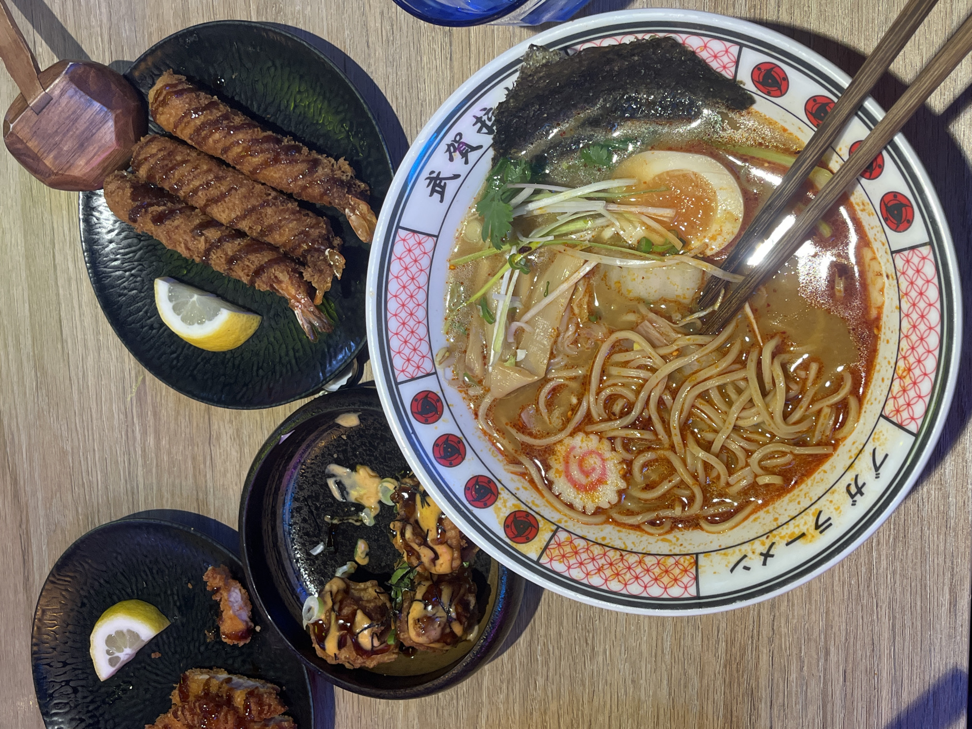 The image shows a top-down view of a table laden with Japanese food. A large bowl of ramen with noodles, broth, an egg, seaweed, and other toppings dominates the scene. Next to it is a black plate containing fried shrimp drizzled with sauce, accompanied by a lemon wedge. Another black plate holds what appear to be fried chicken pieces with lemon, and a third small bowl is filled with other fried items and sauce. Chopsticks rest atop the ramen bowl. The table has a light wood grain finish. It is not possible to discern the specific location within Barcelona, Spain based on the image alone.